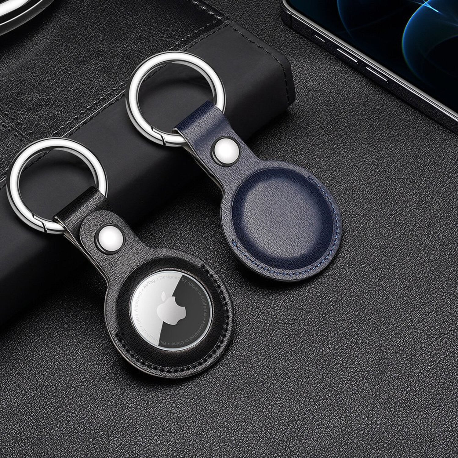 Zwei Apple AirTags auf dunkler Oberfläche. Einer ist schwarz, der andere blau, mit Schlüsselanhängern.