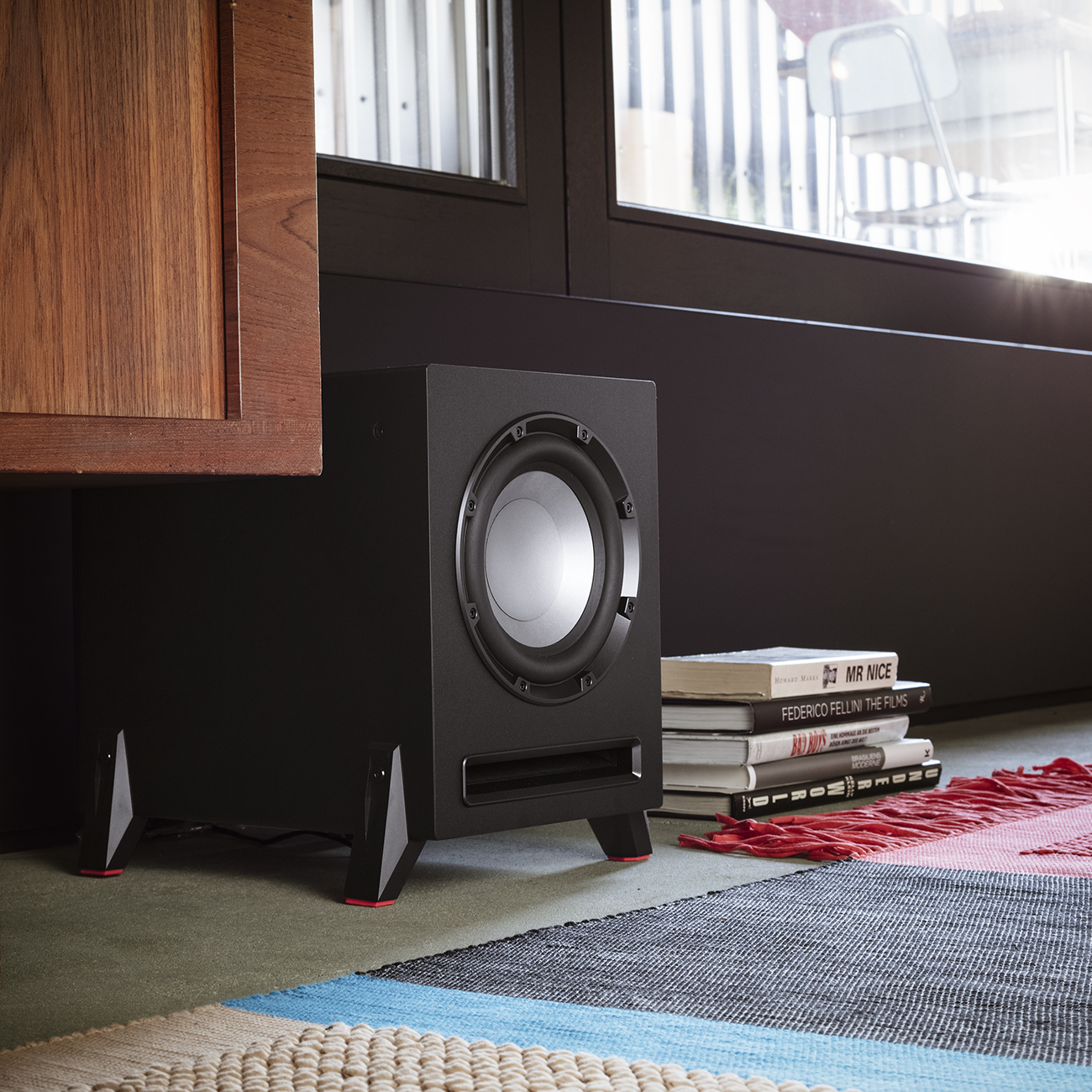 Ein schwarzer Subwoofer mit silbernem Lautsprecher. Er steht auf Beinen mit roten Akzenten, in der Nähe von Büchern und einem bunten Teppich.