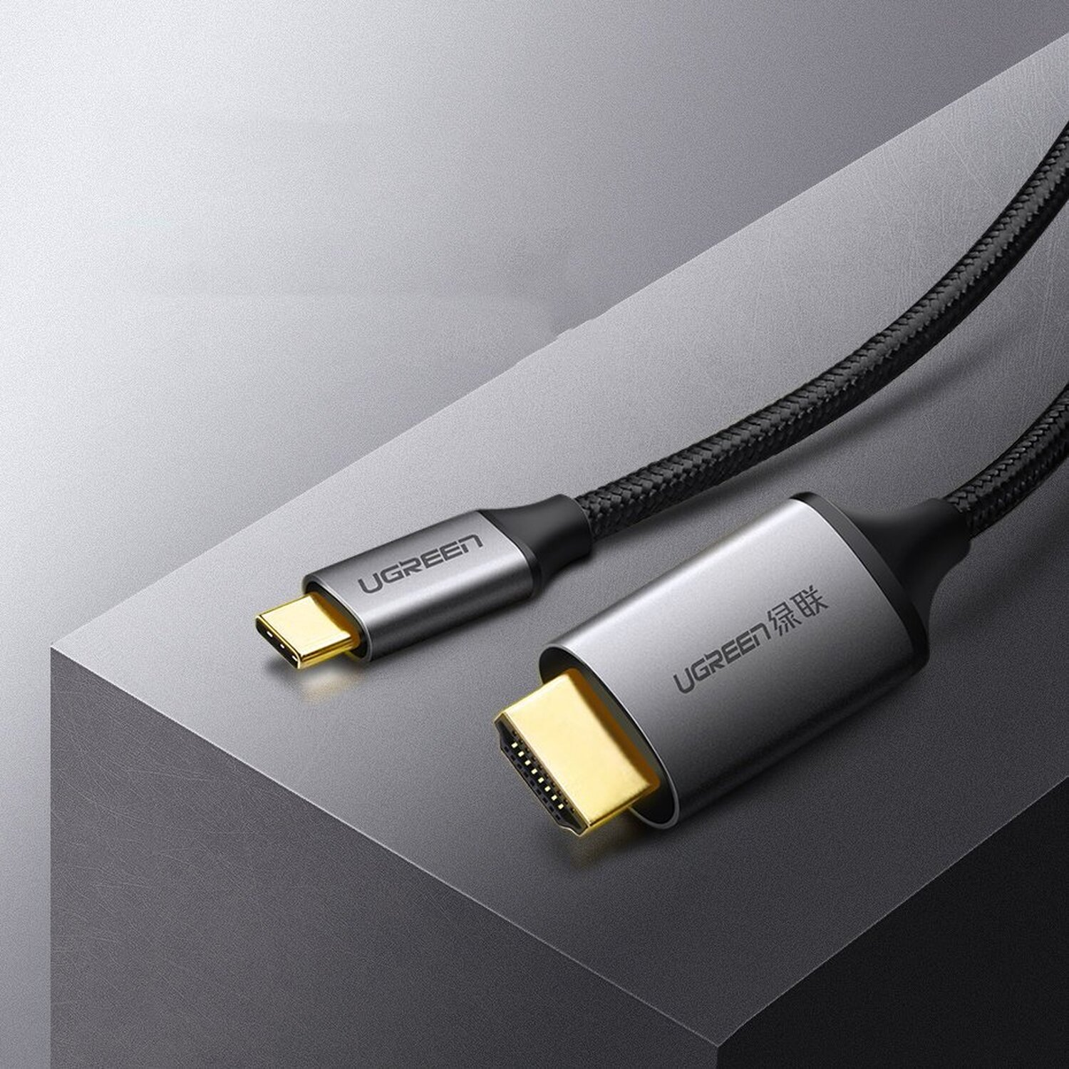 Dwa kable UGREEN. Jeden USB-C i jeden HDMI, oba srebrne z czarnymi plecionymi kablami, na szarej powierzchni.