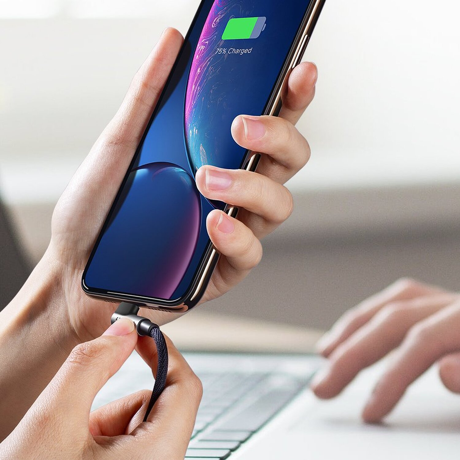 Hand hält Handy mit Ladekabel. Eine andere Hand liegt auf einem Schreibtisch.