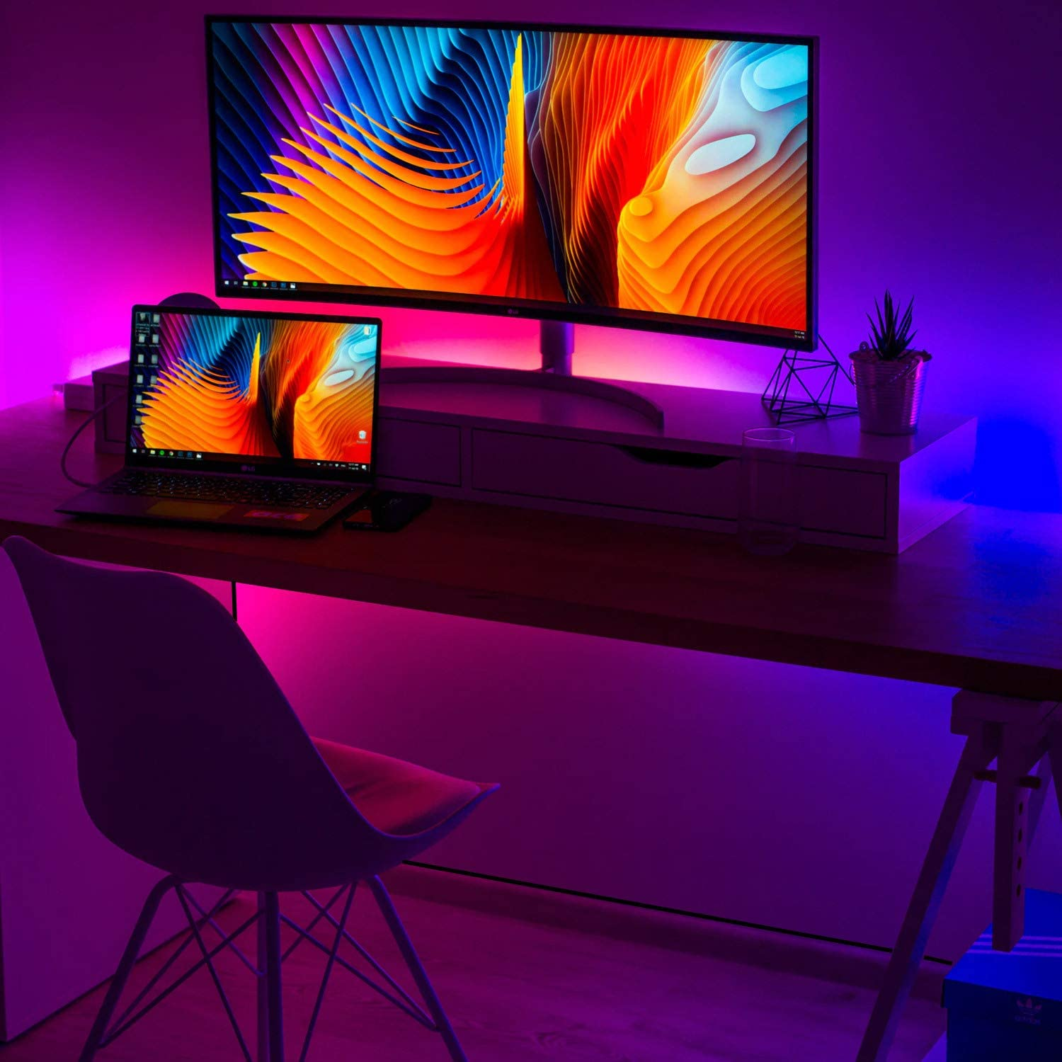 Schreibtisch mit Monitor, Laptop und Pflanze. Pinke und blaue Neonlichter beleuchten die Szene.