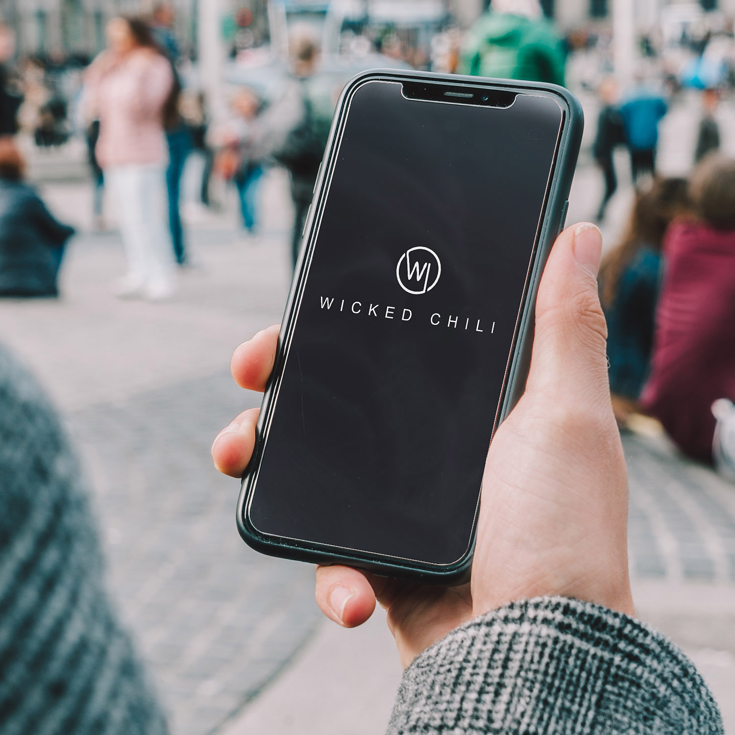 Eine Person hält ein Telefon mit dem Wicked Chili Logo auf dem Bildschirm, in einer unscharfen Umgebung im Freien.