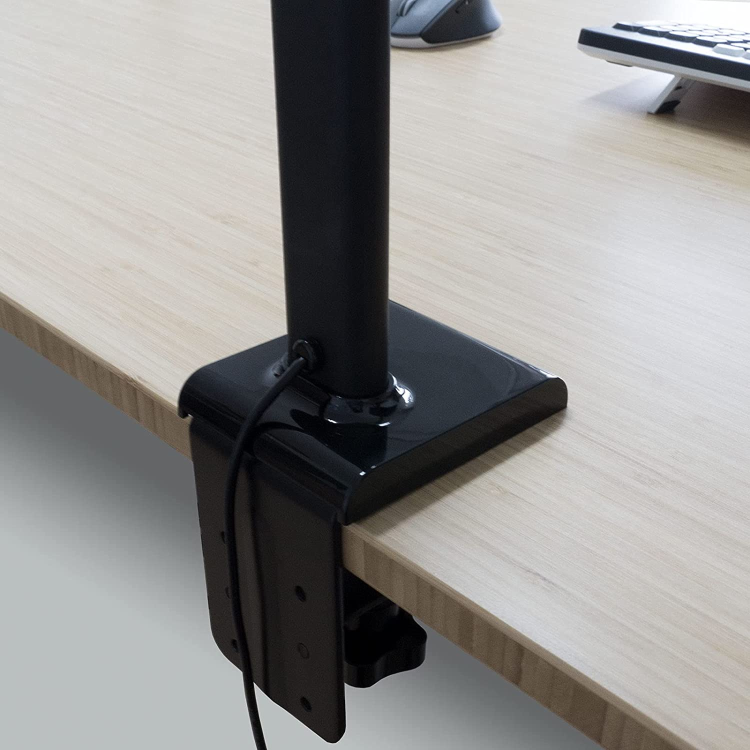 Schwarzer Monitorständer an einem hellen Holzschreibtisch befestigt. Schwarzes Kabel sichtbar.