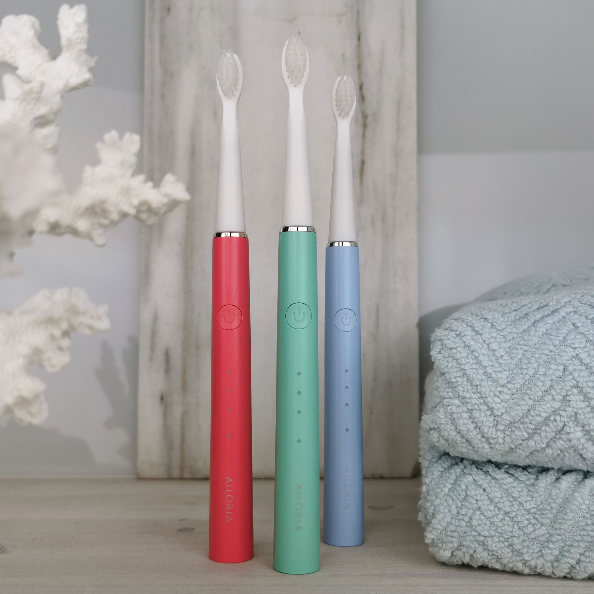 Tres cepillos de dientes eléctricos: rojo, verde y azul, sobre madera con una toalla.