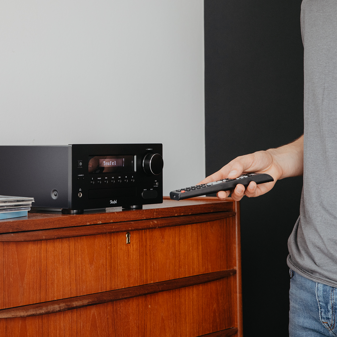 Die Hand eines Mannes in der Nähe eines schwarzen Stereo-Systems, die eine Fernbedienung hält. Das System steht auf einem Holzschrank.