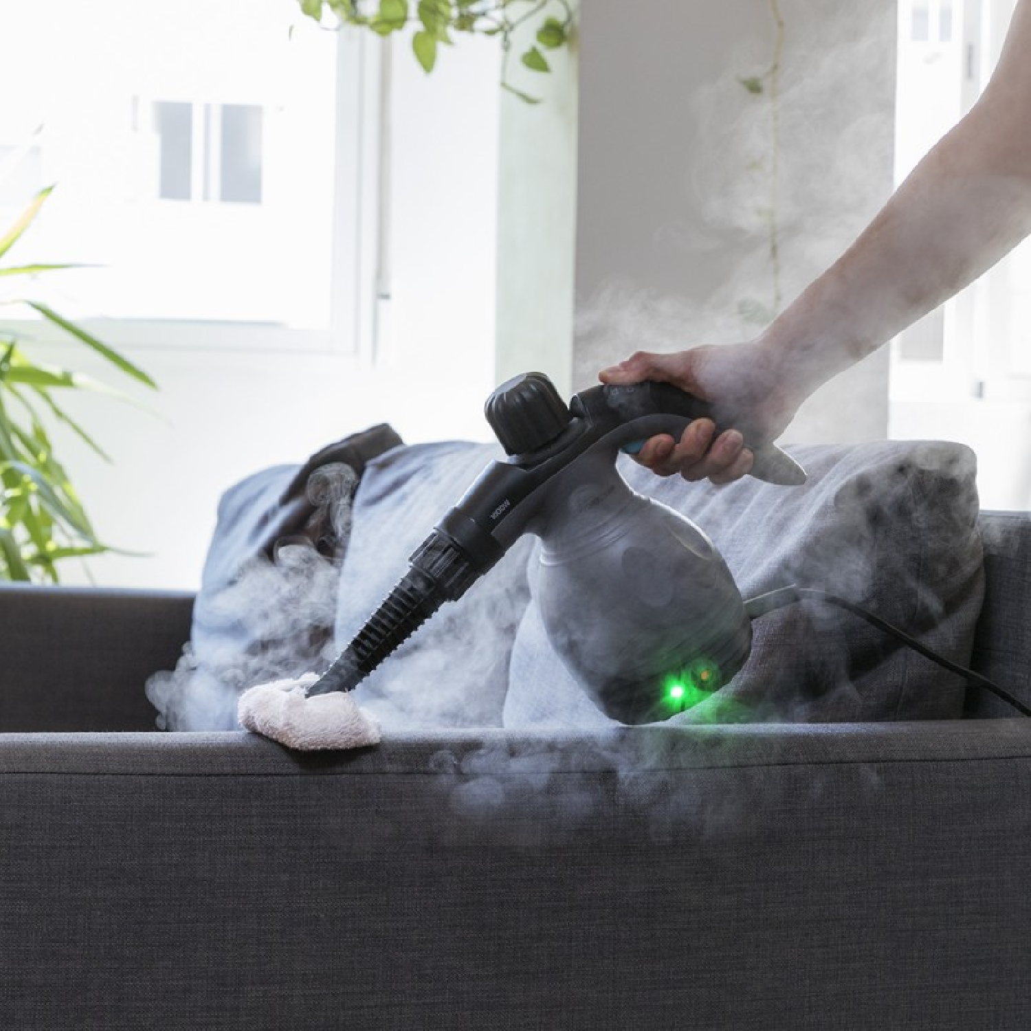 Eine Person benutzt einen Dampfreiniger auf einem grauen Sofa. Dampf steigt aus der Düse auf.