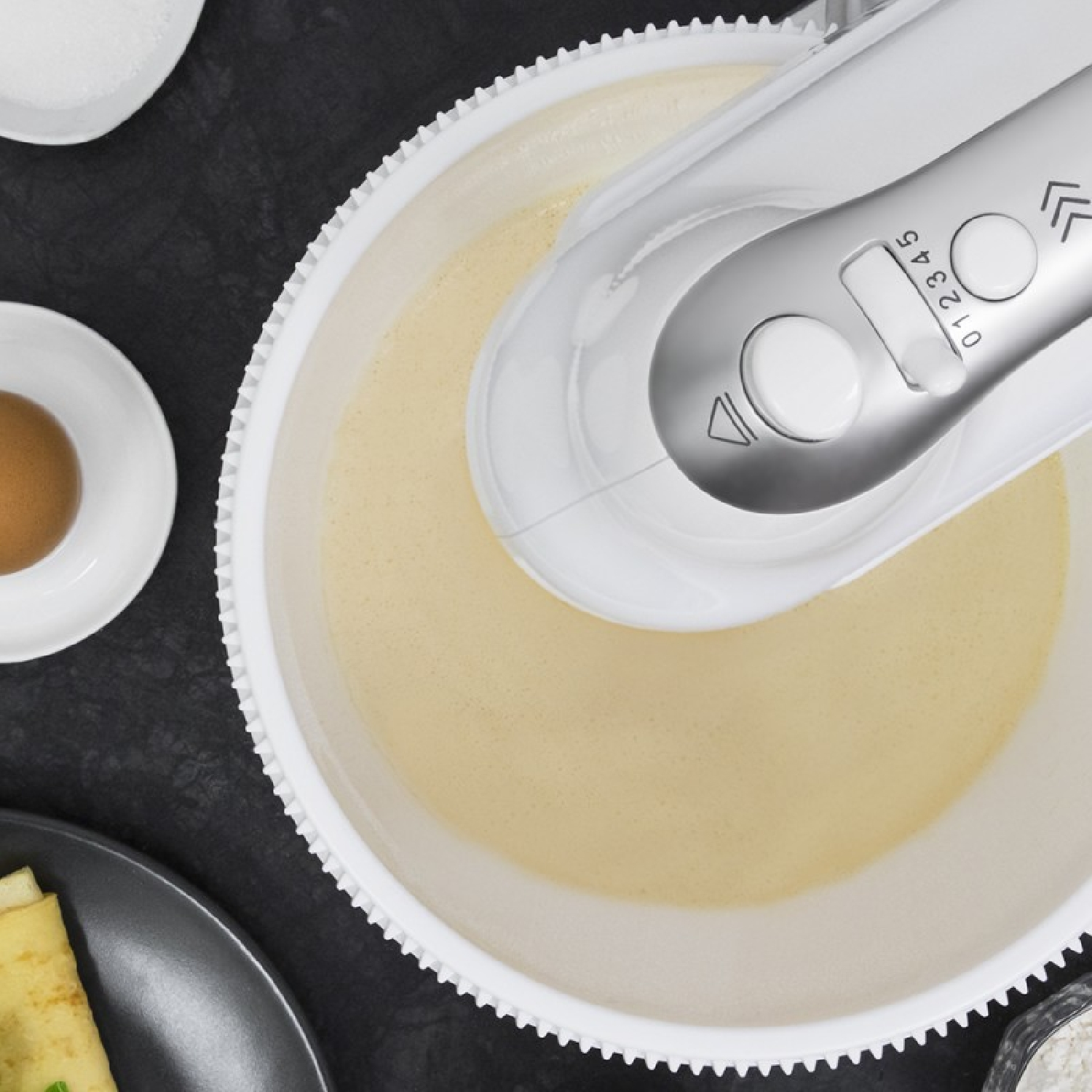 Una batidora mezclando una masa en un cuenco blanco. Los ingredientes están al lado. Vista cenital.