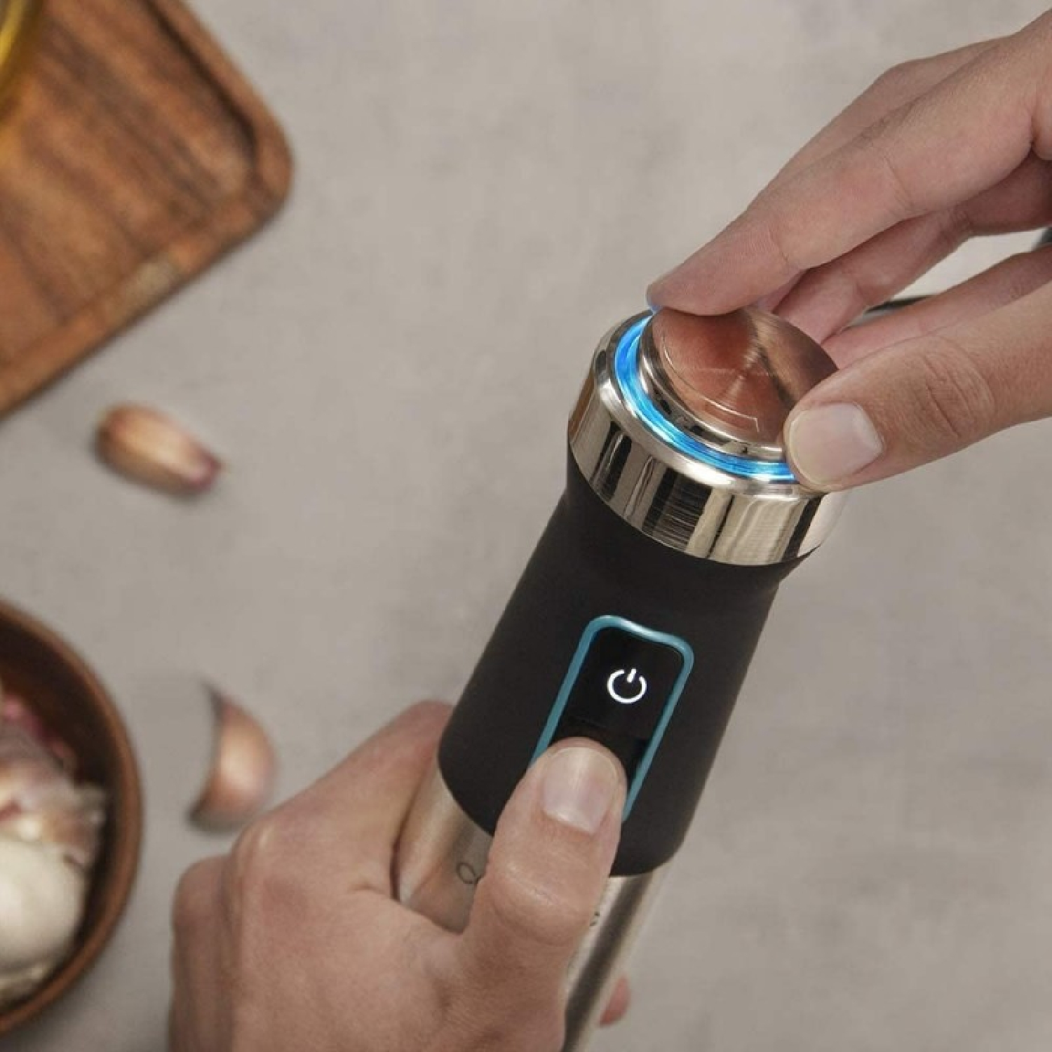 Een hand drukt op de aan/uit-knop van een zwart-zilveren handheld staafmixer met blauw licht.