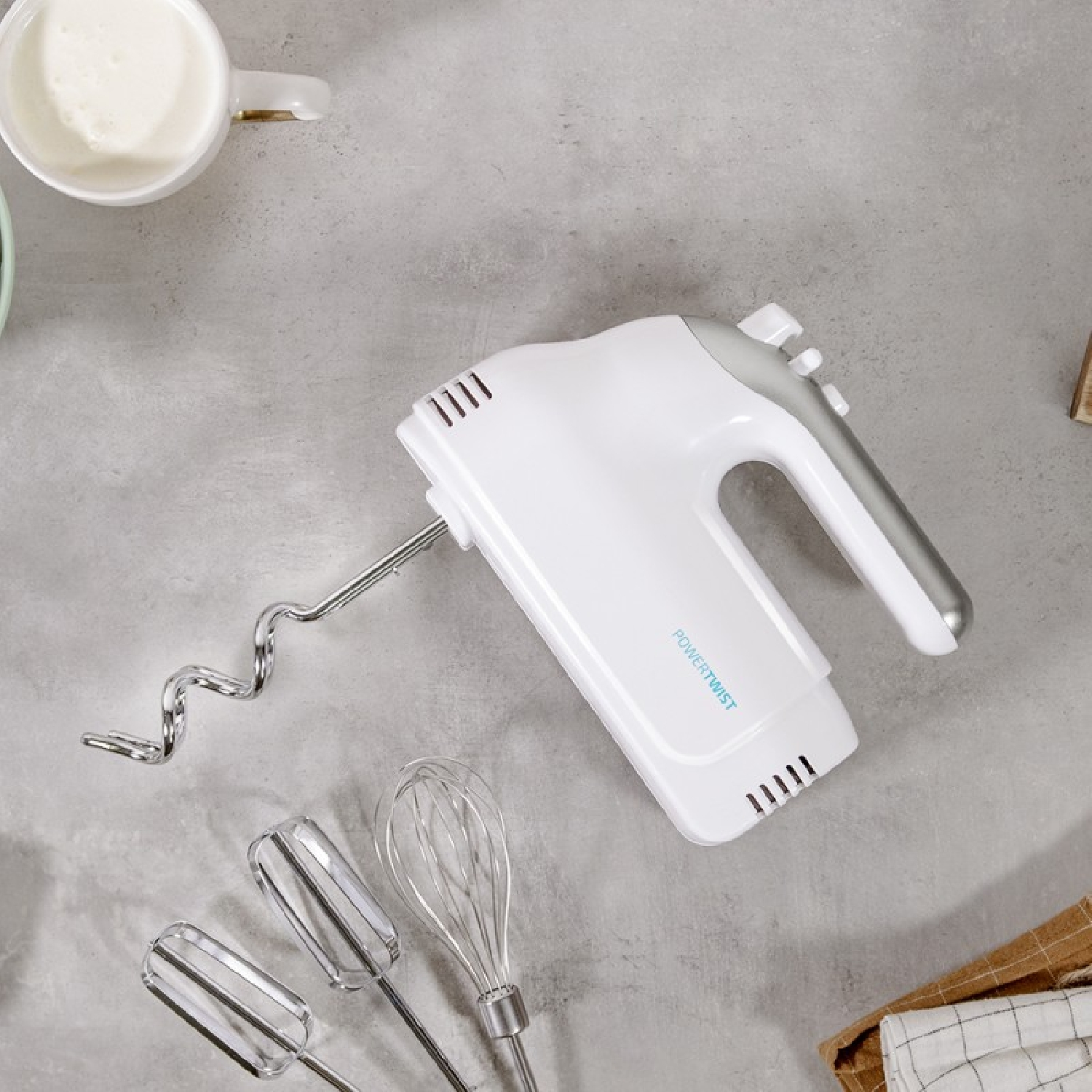 Weißer Handmixer mit Aufsätzen auf grauem Untergrund, mit Tasse und Handtuch.