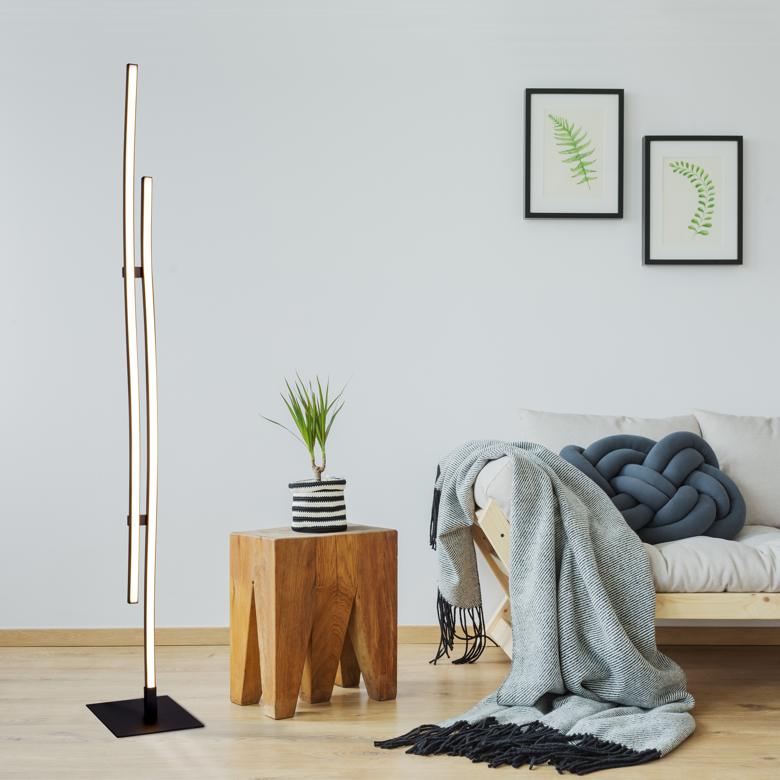 Moderne Stehlampe mit drei Lichtbalken und einem Holzschemel mit Pflanze, sowie ein Sofa mit Decke.