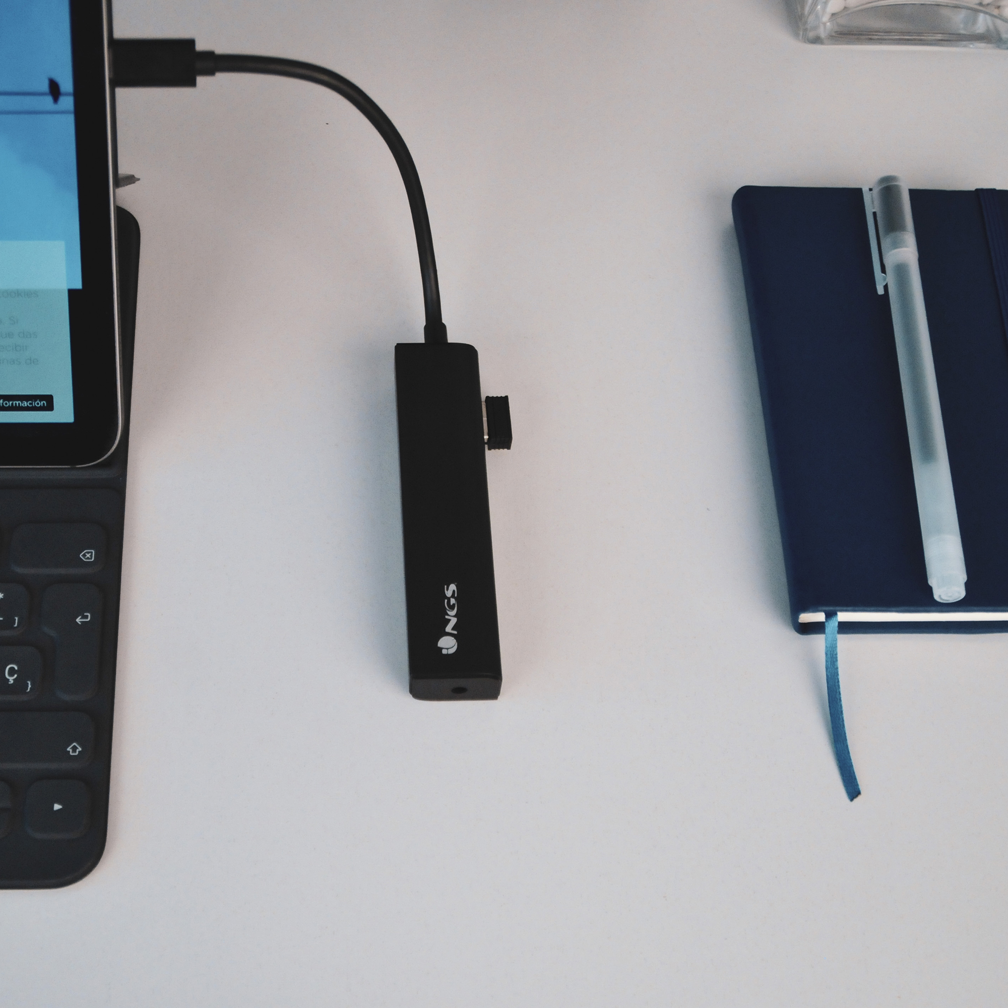 Tableta con teclado y un adaptador negro conectado. Un cuaderno y un bolígrafo al lado.
