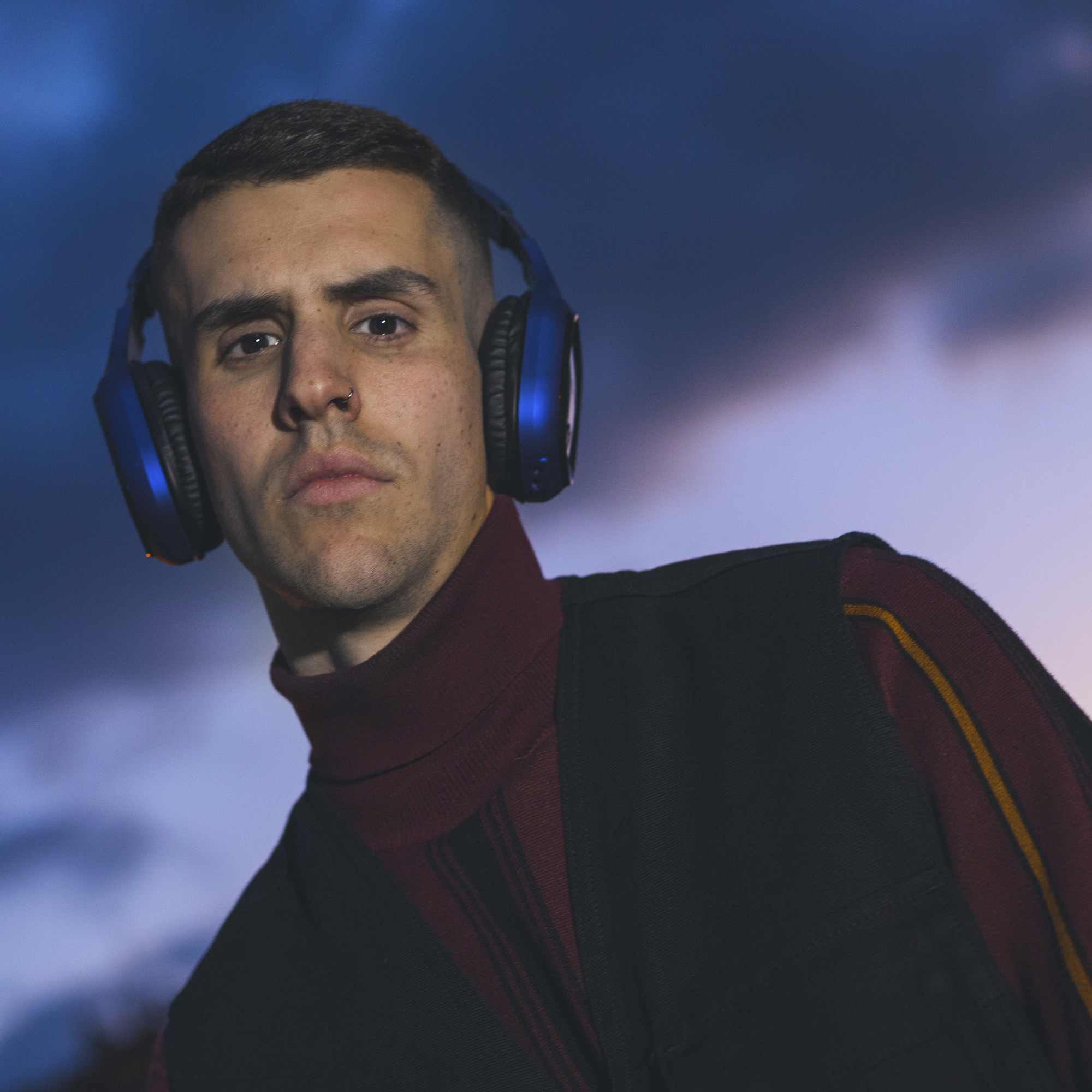 Uomo con cuffie blu, gilet nero e camicia rossa. Lo sfondo è un cielo blu nuvoloso.