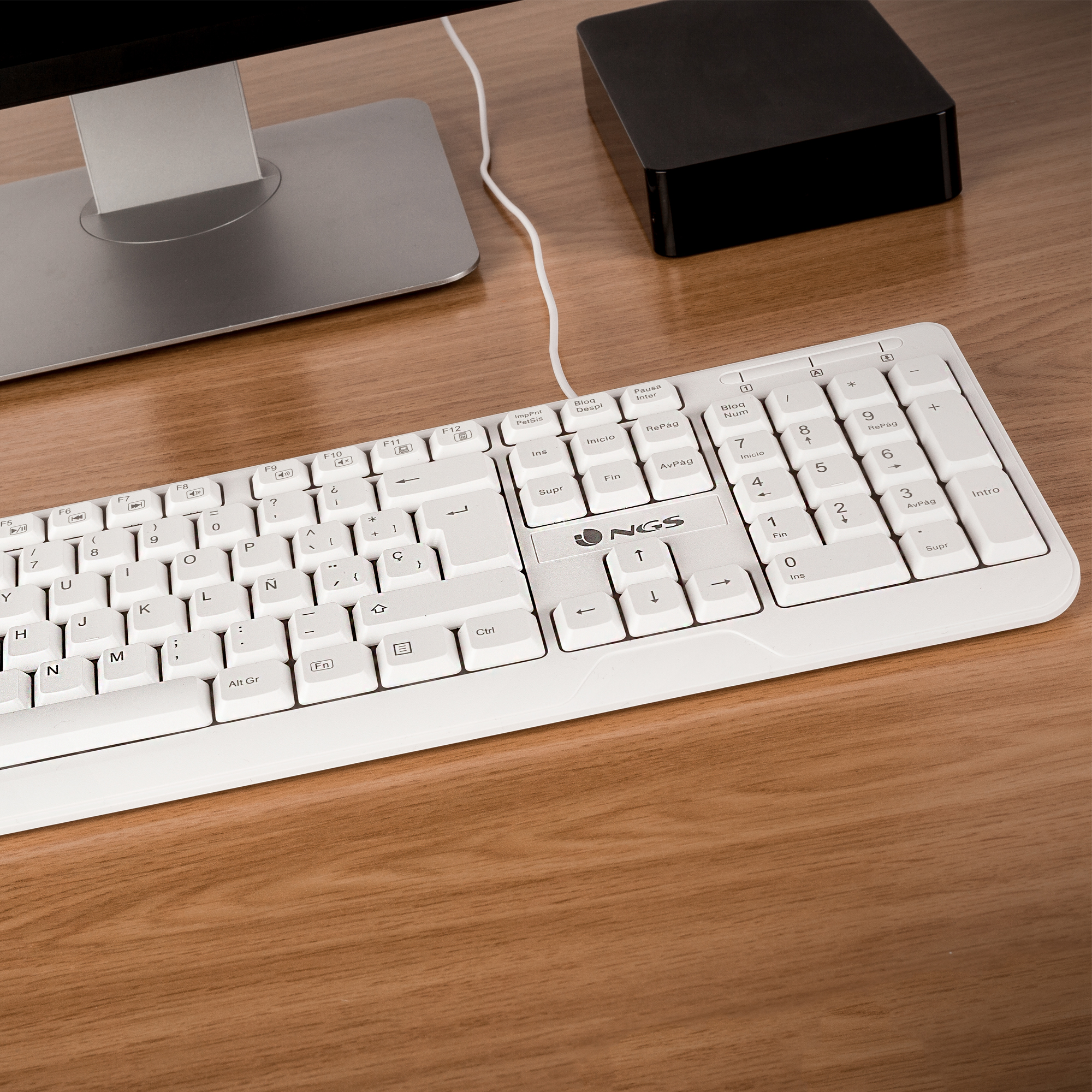 Weiße Computertastatur mit Tasten, auf einem Holzschreibtisch, neben einem Monitor und einem schwarzen Gerät.