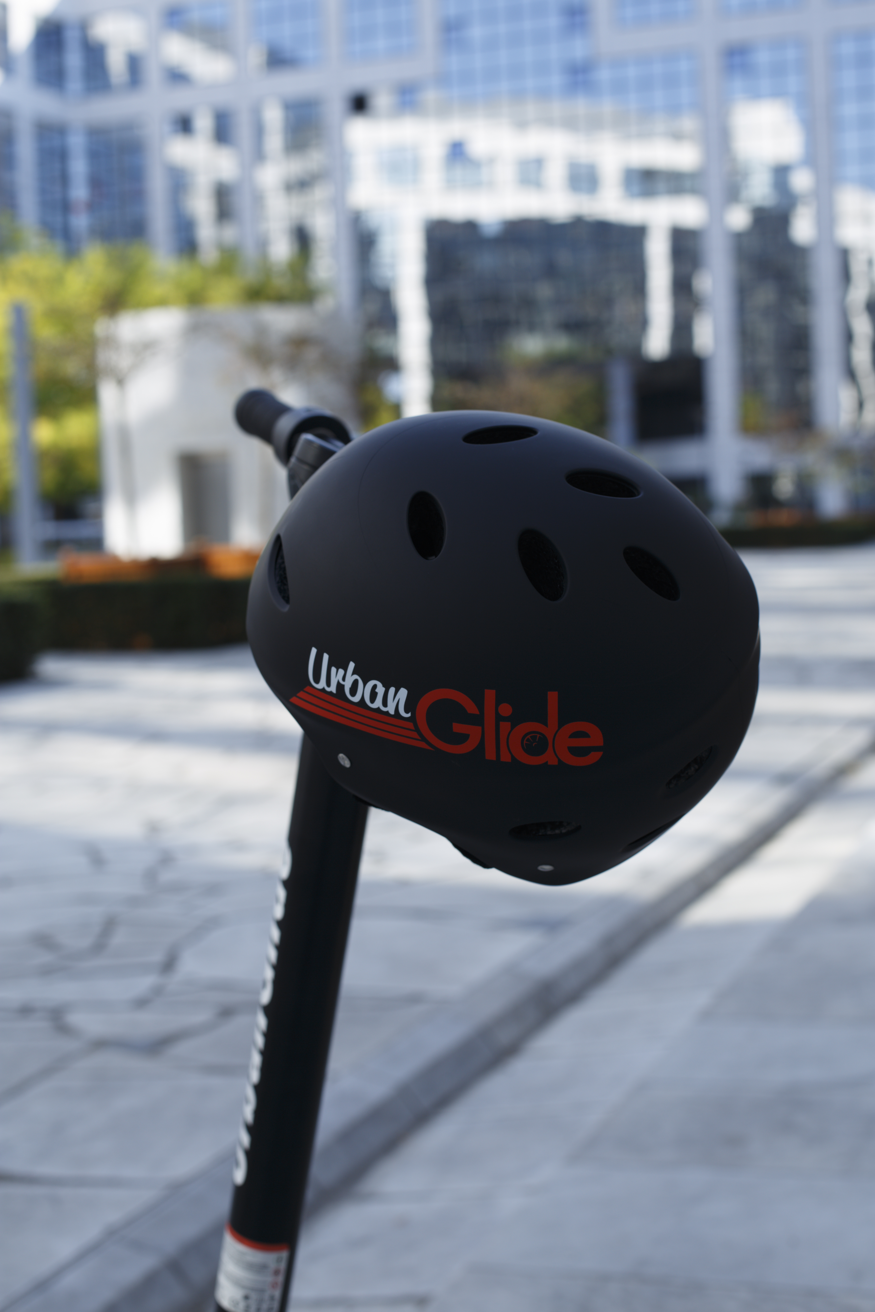 Un casco negro Urban Glide sobre el manillar de un patinete negro. El texto 'Urban Glide' está en el casco.