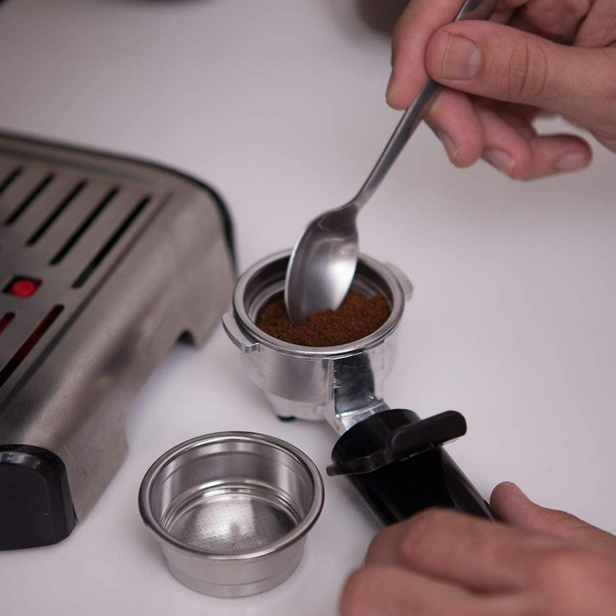 Osoba używająca łyżki do wkładania zmielonej kawy do kolby. W pobliżu ekspres do espresso.