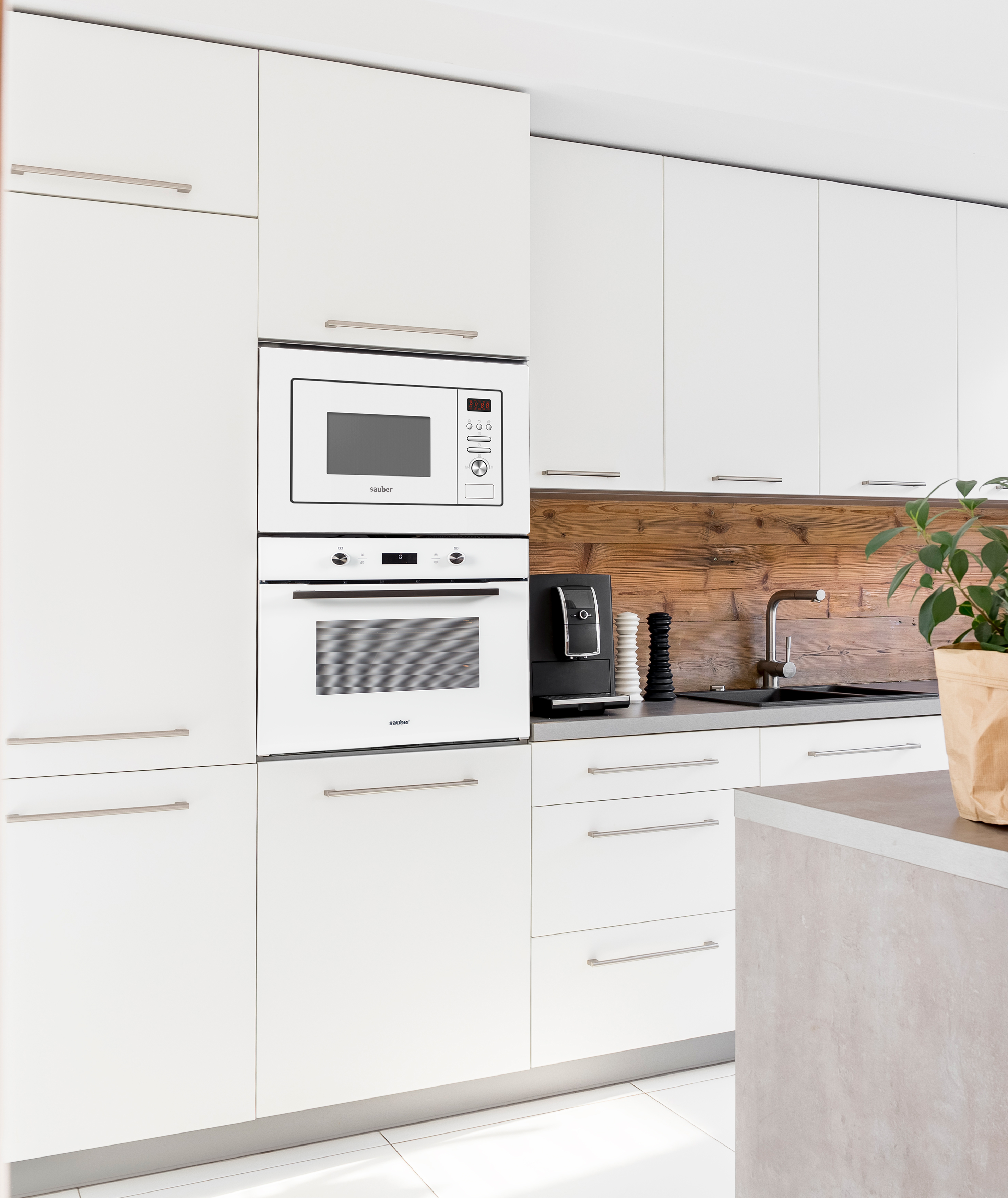 Cocina blanca moderna con gabinetes blancos, microondas, horno y salpicadero de madera. Hay una planta.
