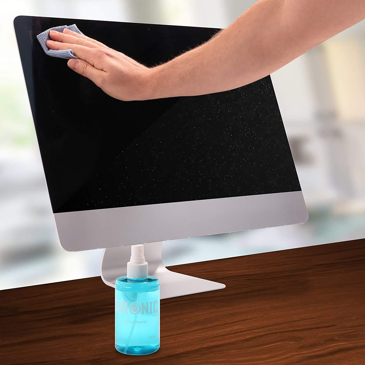 Eine Hand wischt einen Computerbildschirm mit einem Tuch ab. Darunter steht eine Sprühflasche auf einer braunen Oberfläche.