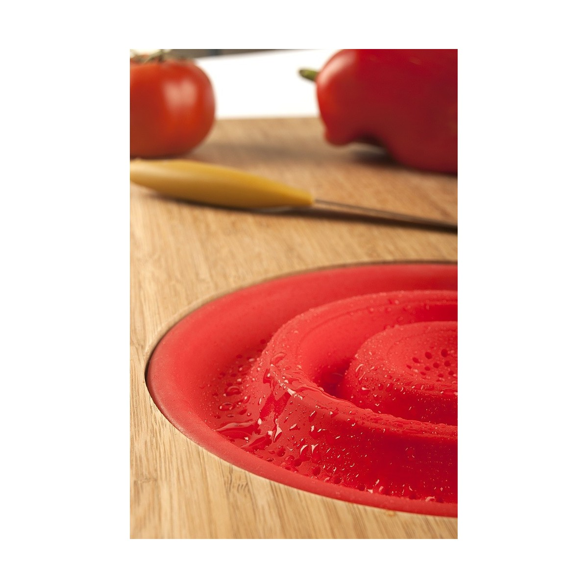 Colador de silicona rojo en tabla de cortar de madera, con cuchillo y verduras, primer plano.
