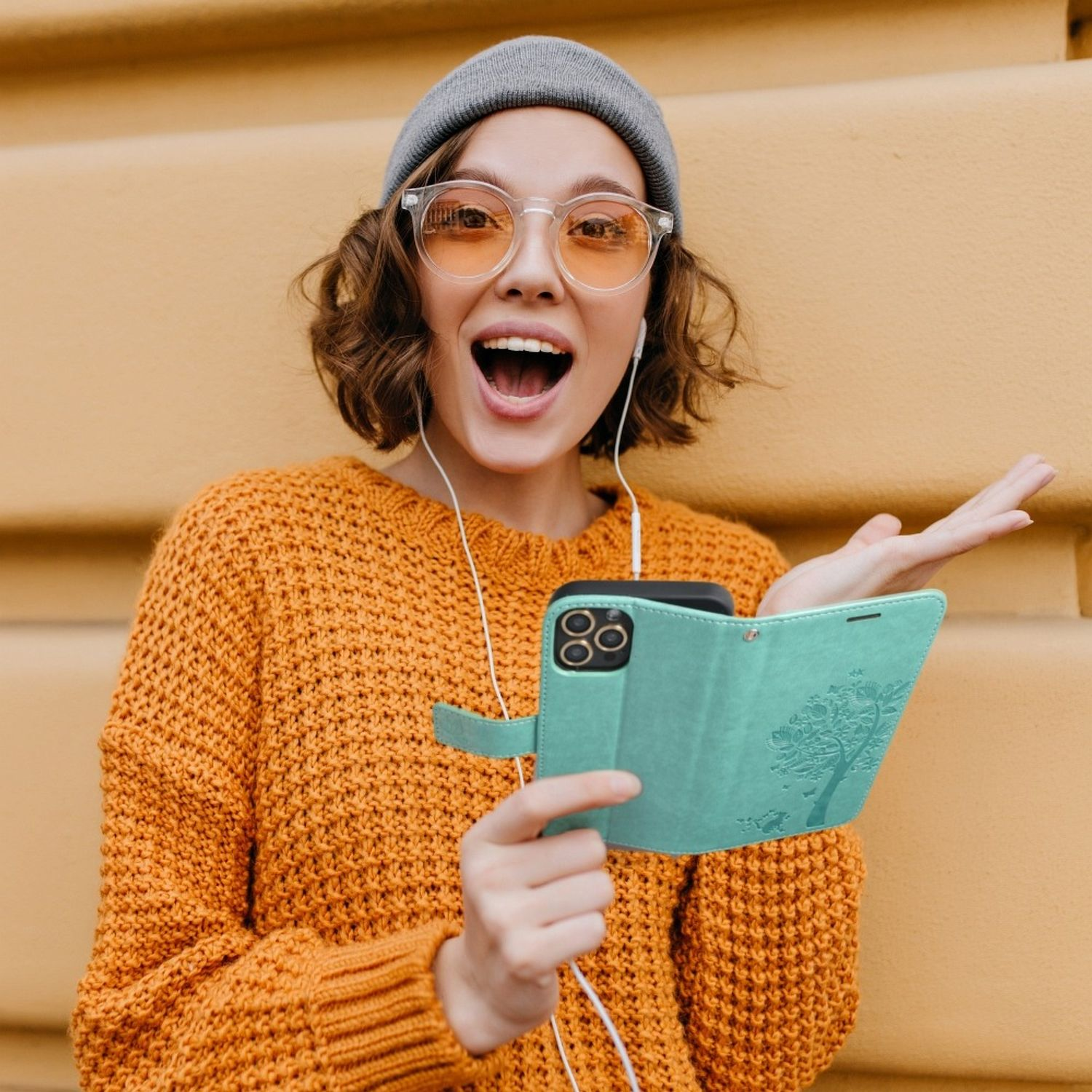 Frau mit Brille und orangefarbenem Pullover, die eine türkisblaue Handyhülle mit Baumdesign hält.