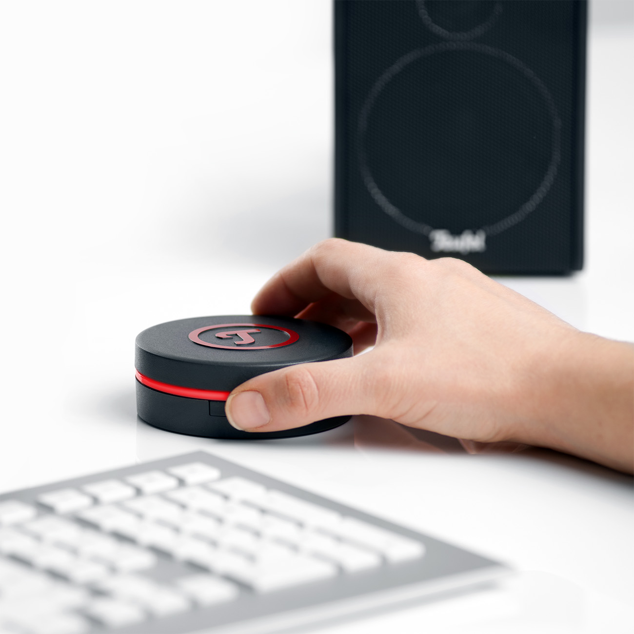 Eine Hand hält einen schwarz-roten Knopf. Ein Lautsprecher und eine Tastatur sind im Hintergrund.