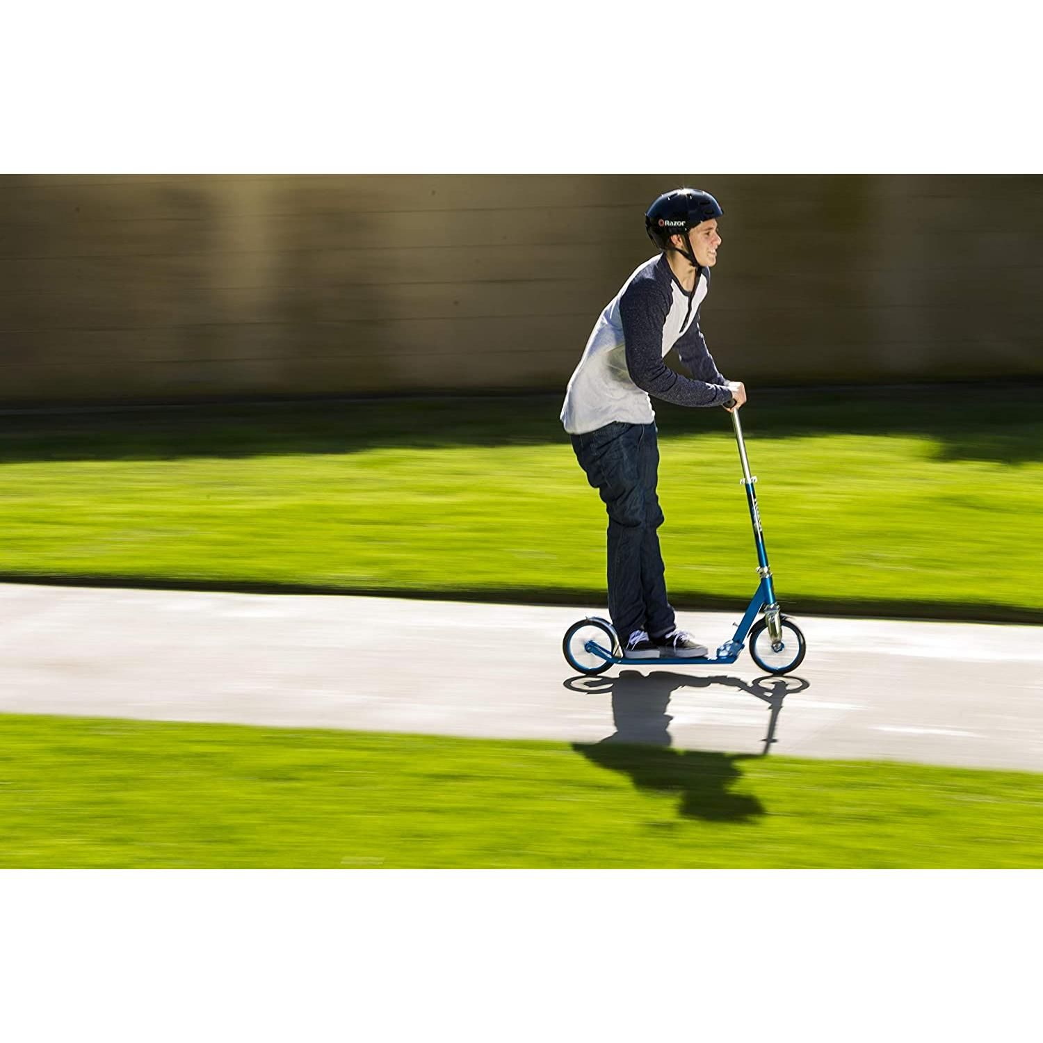 Une personne sur une trottinette bleue sur un trottoir, herbe et clôture.