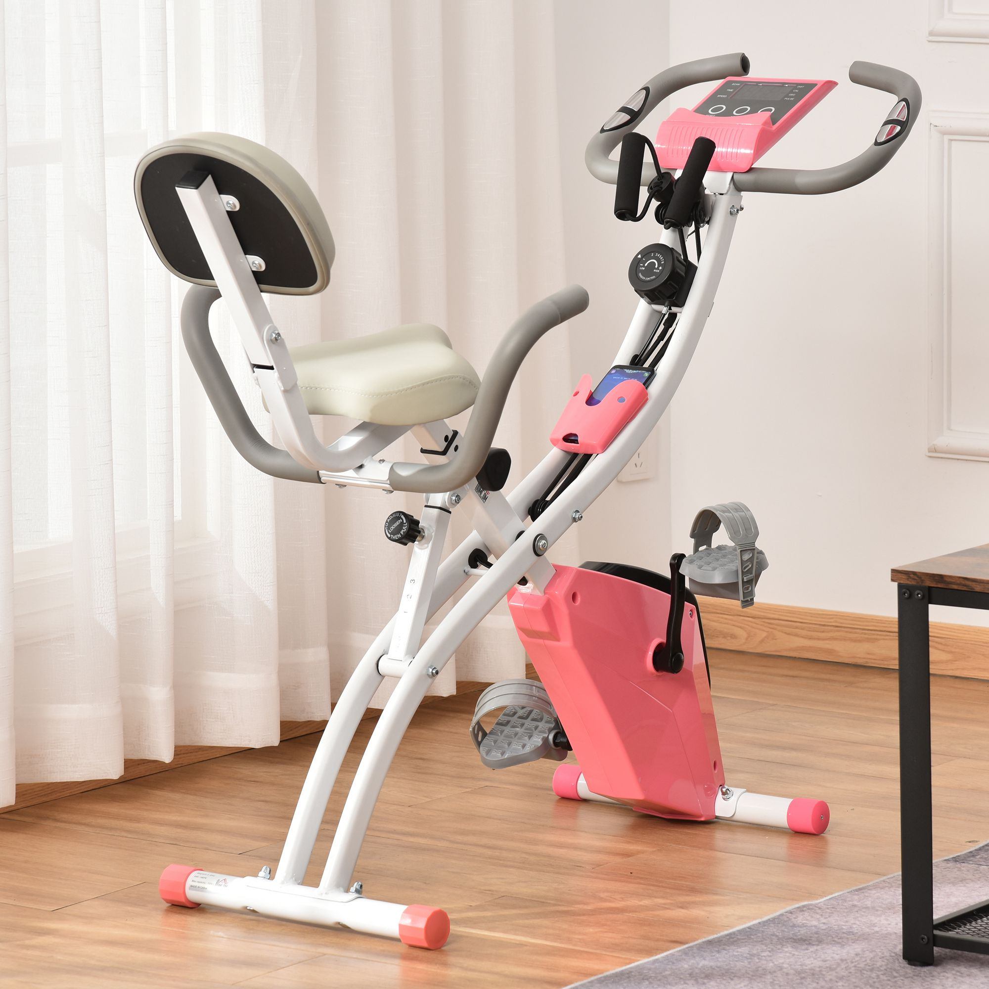 Ein pink-weißer Heimtrainer steht auf einem Holzboden vor einem hellen Vorhang.