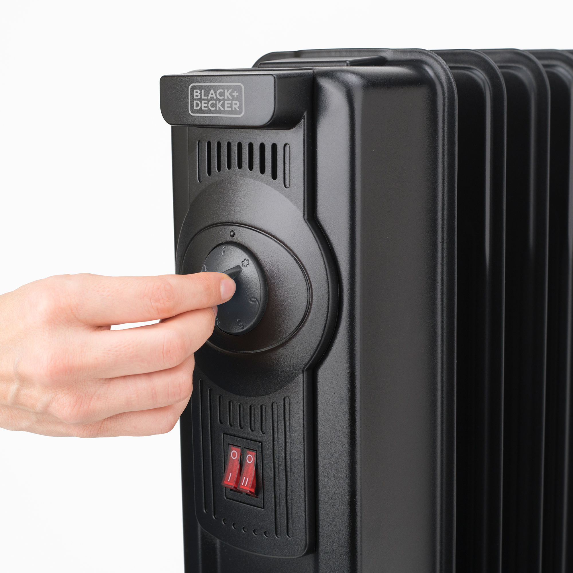 Ręka obraca pokrętło na czarno-srebrnym grzejniku olejowym, widoczne czerwone przełączniki zasilania.