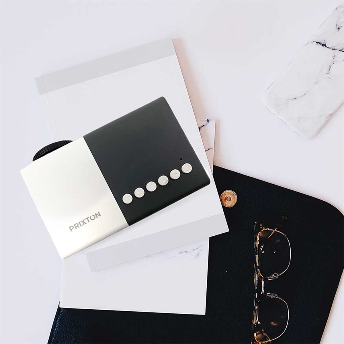A black and white projector, a black purse, notebooks, and glasses on a white surface.