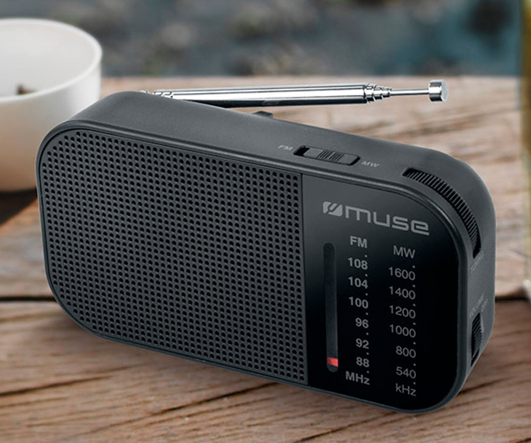 Czarny przenośny radio z logo Muse, stojący na drewnianym stole z białym kubkiem.