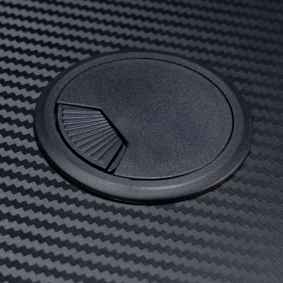 Pasacables negro sobre superficie con patrón de fibra de carbono. El pasacables es circular y ranurado.