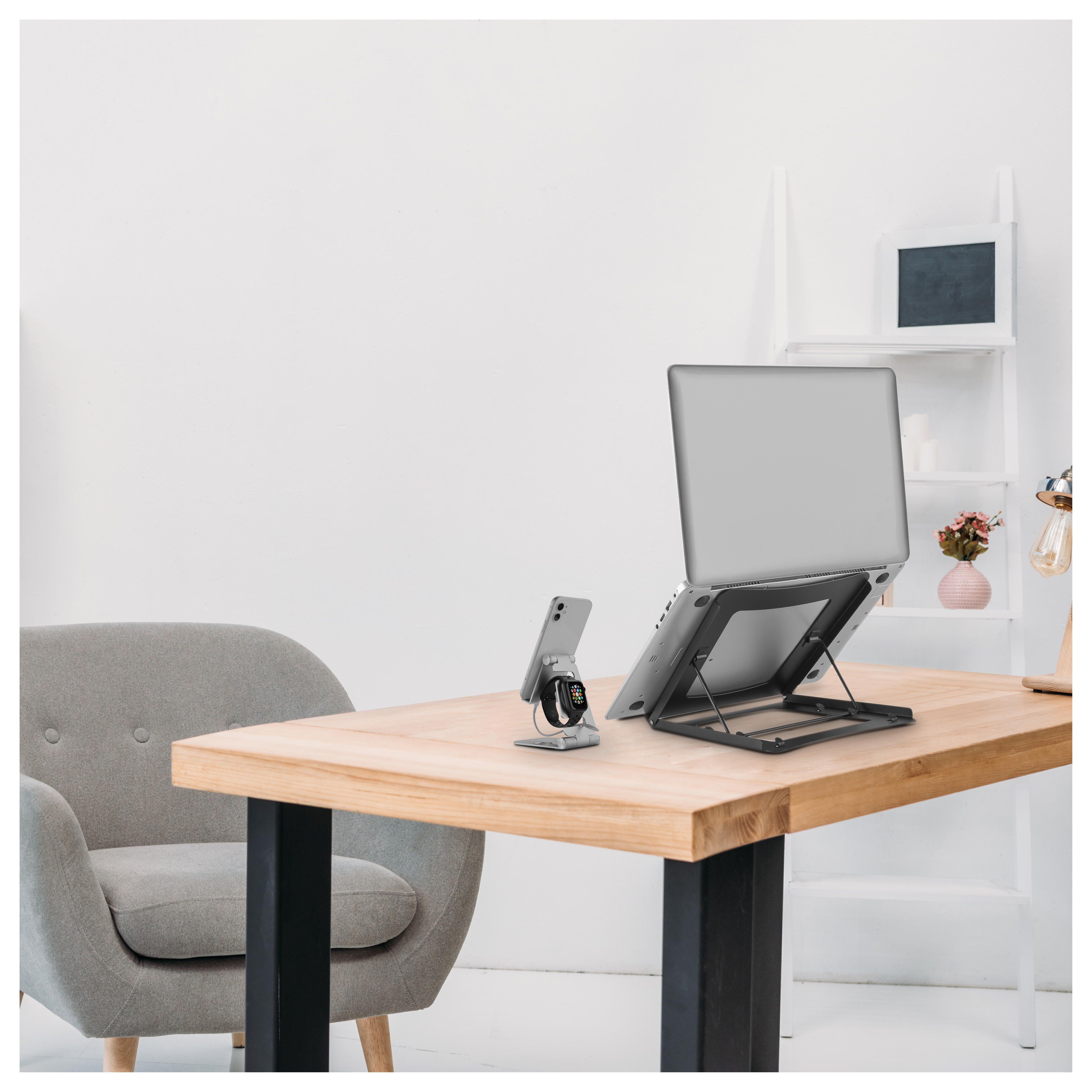 Laptop en soporte, teléfono y smartwatch en un escritorio. Silla gris y pared blanca al fondo.