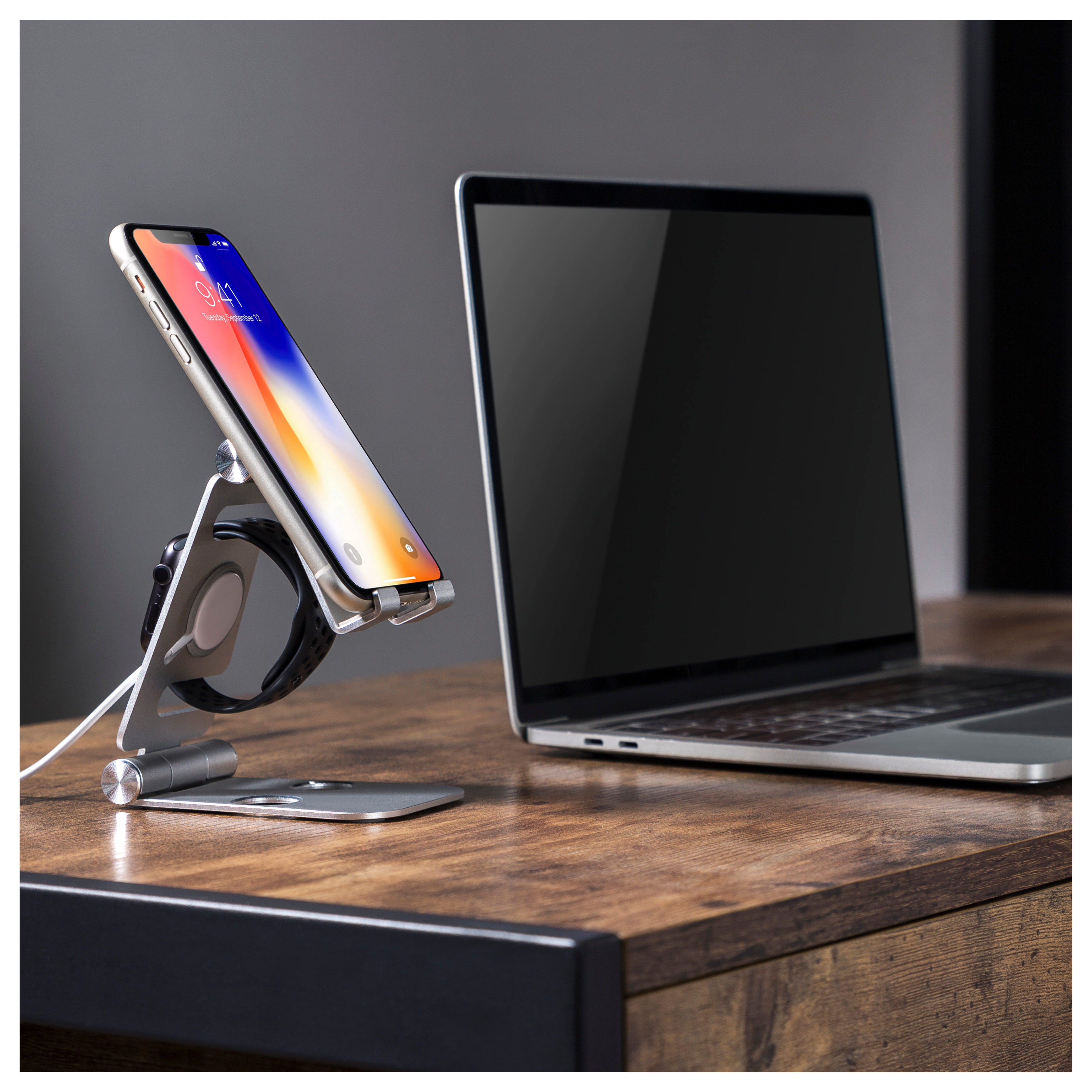 Smartphone en un soporte plateado, junto a un portátil en un escritorio de madera.