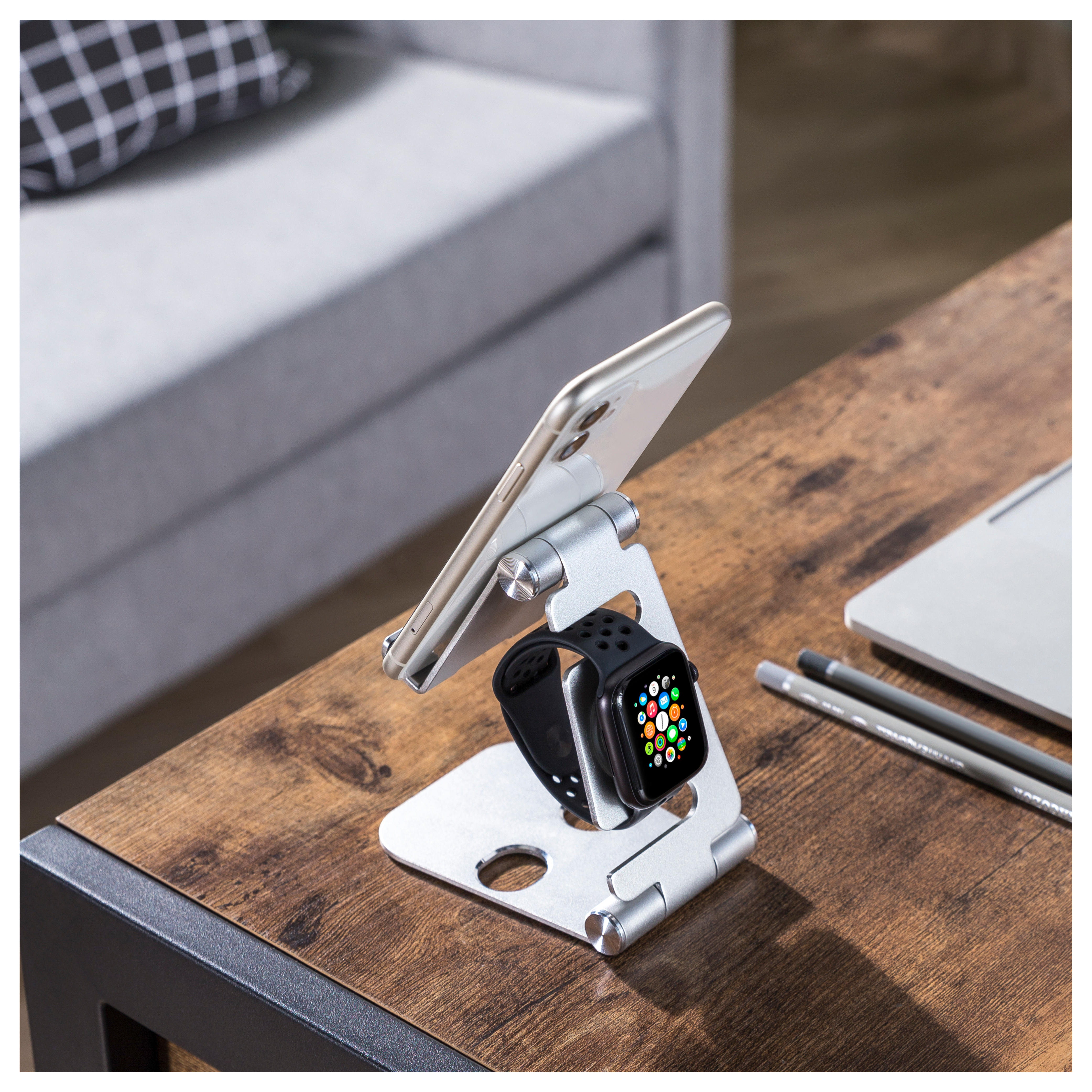 Teléfono y reloj inteligente en un soporte. Lápices en mesa de madera. Sofá gris detrás.