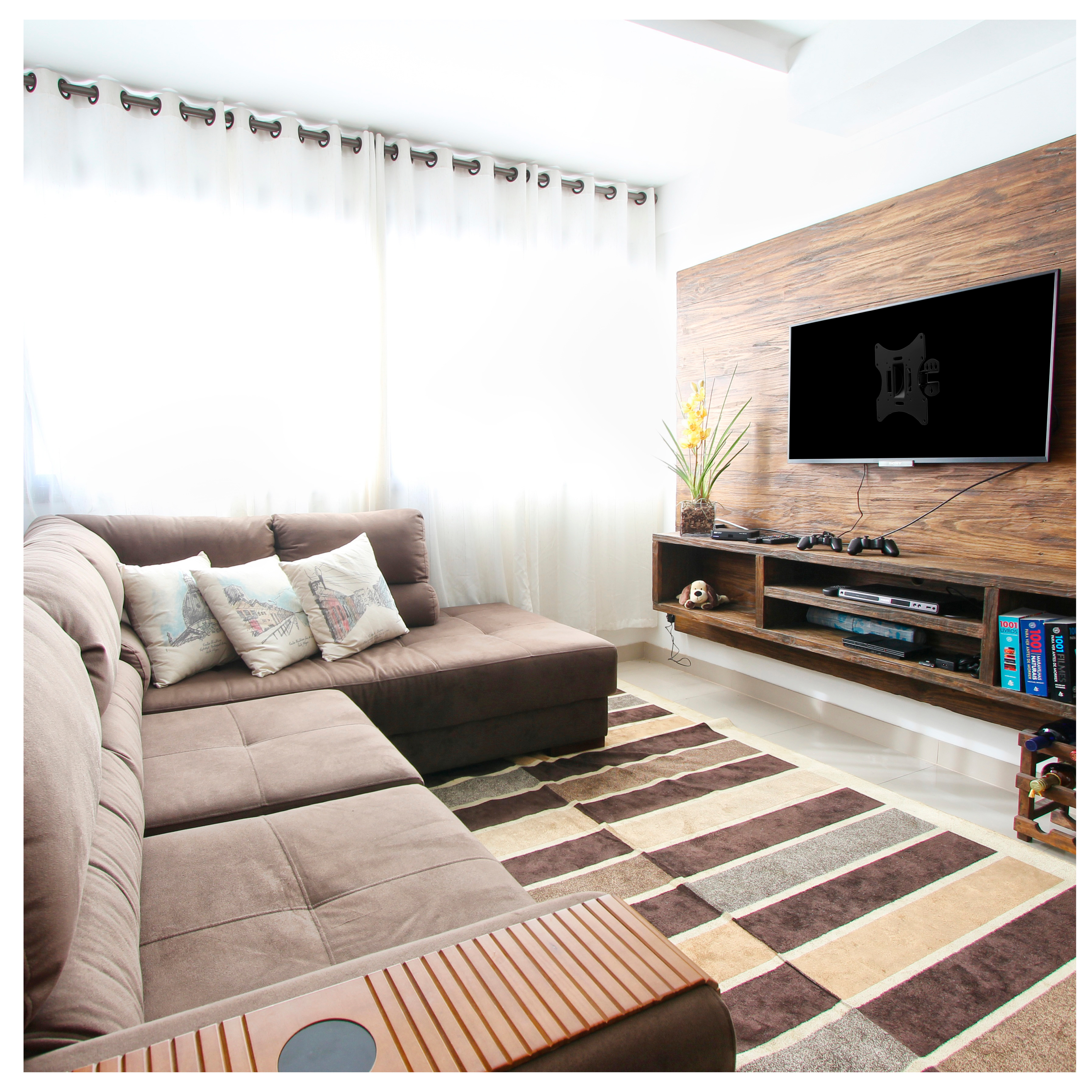 Salón con sofá seccional marrón, TV en pared de madera. Se ve una alfombra y cortinas.