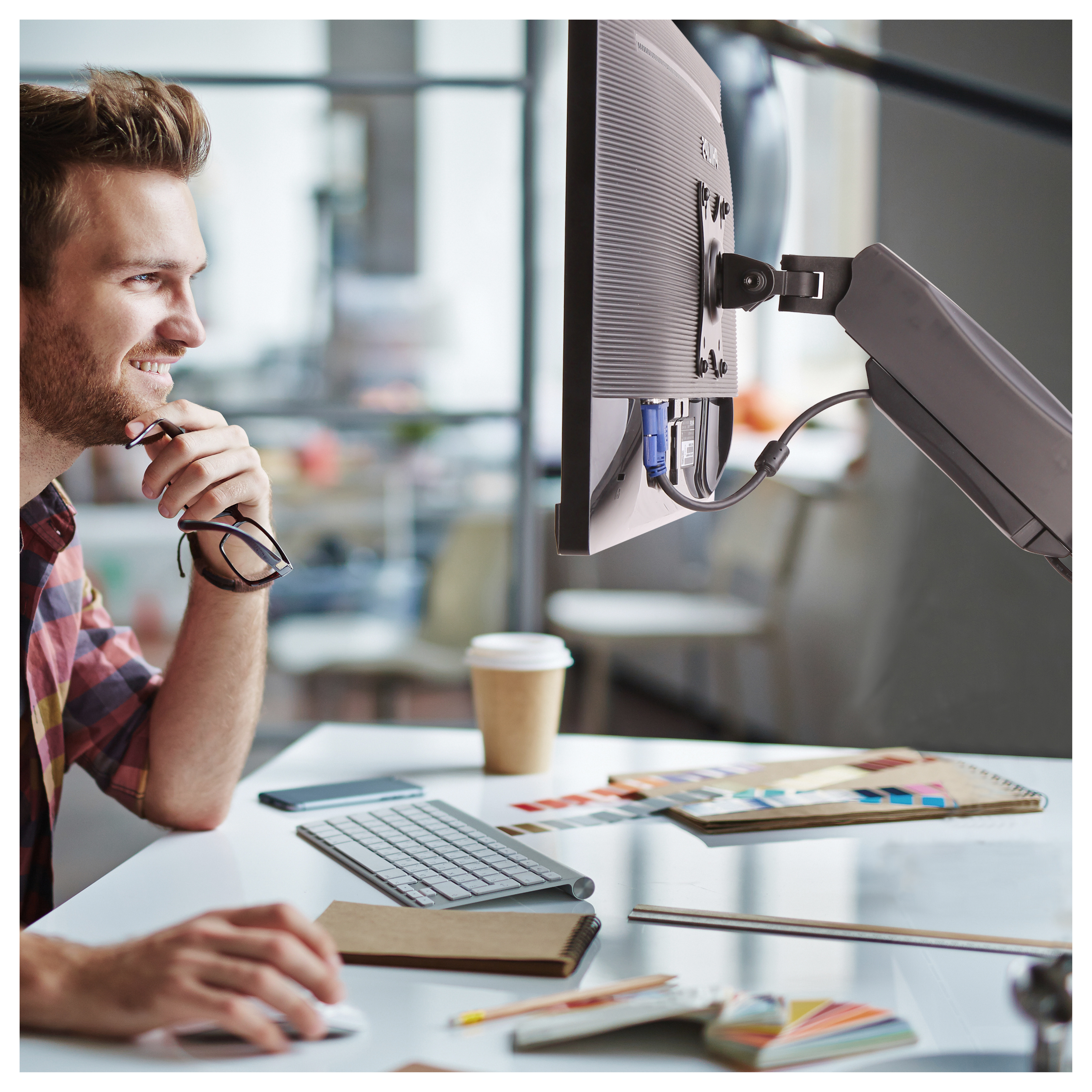 Un hombre mirando un monitor de computadora con un brazo de monitor, junto a una taza de café y un teclado.
