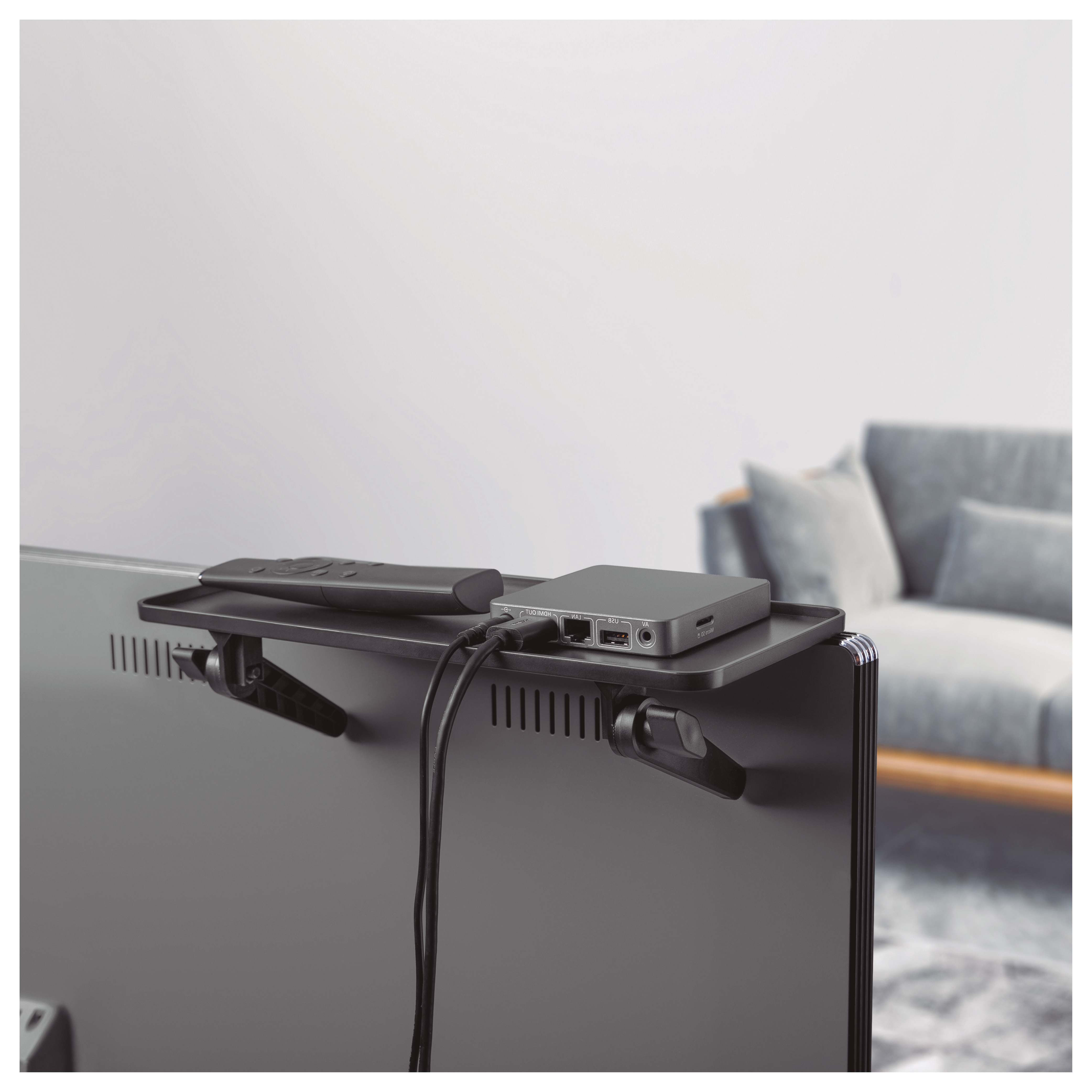 Estante negro en la parte trasera de un televisor, con mando, caja y cables, fondo de sofá gris.