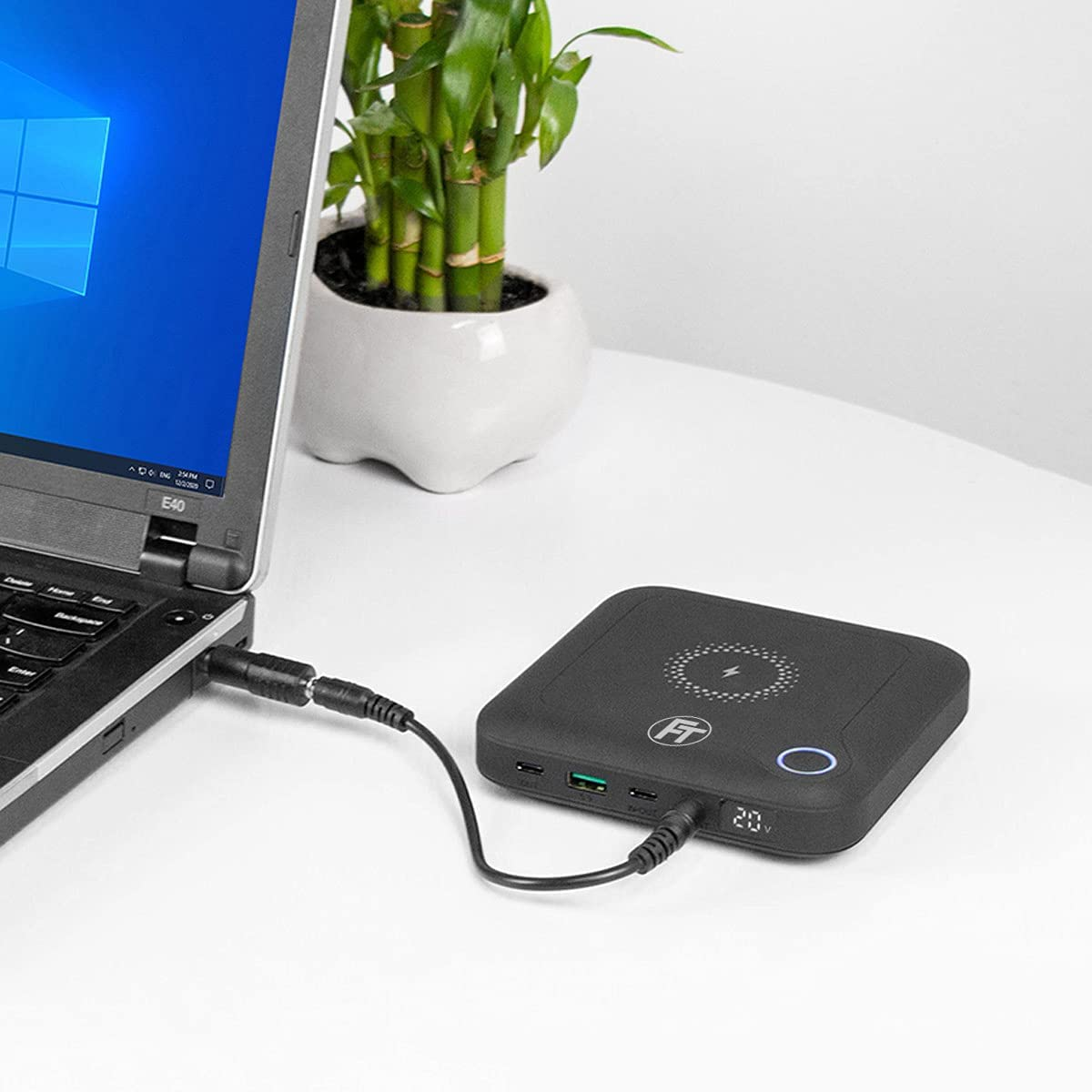 Ein Laptop, der mit einer schwarzen, quadratischen Powerbank verbunden ist. Eine grüne Pflanze ist im Hintergrund.