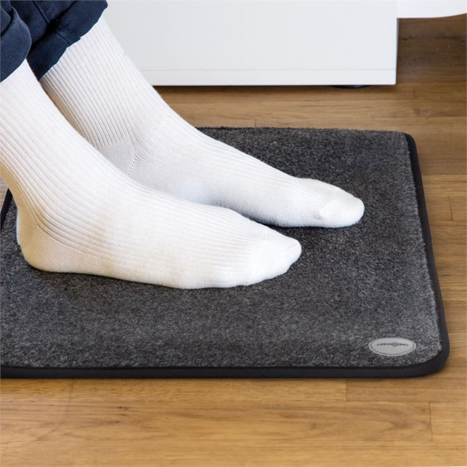 Füße einer Person in weißen Socken auf einem grauen Fußwärmer auf Holzboden.