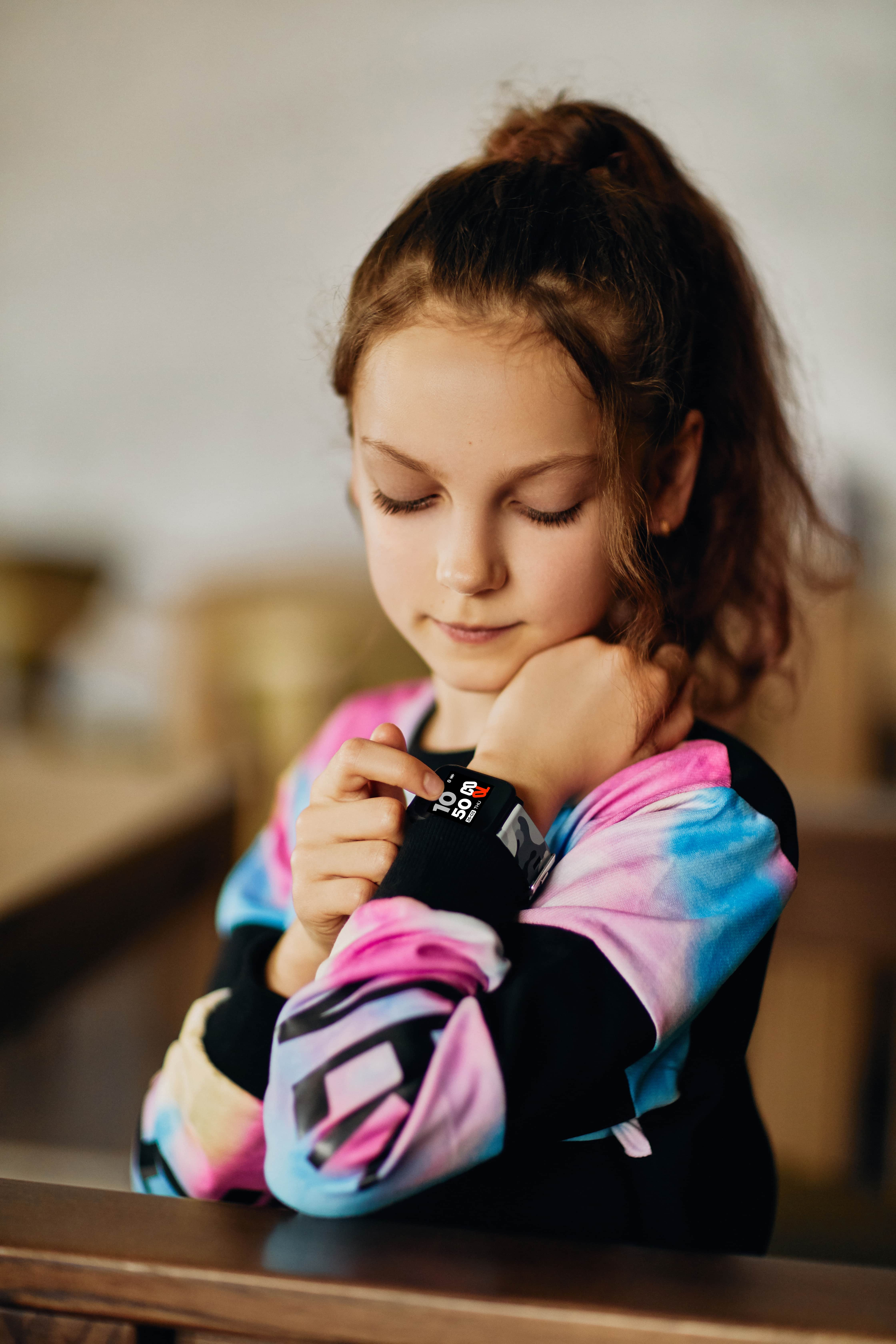 Una niña con un reloj inteligente, mirándolo, sentada. El fondo está borroso.