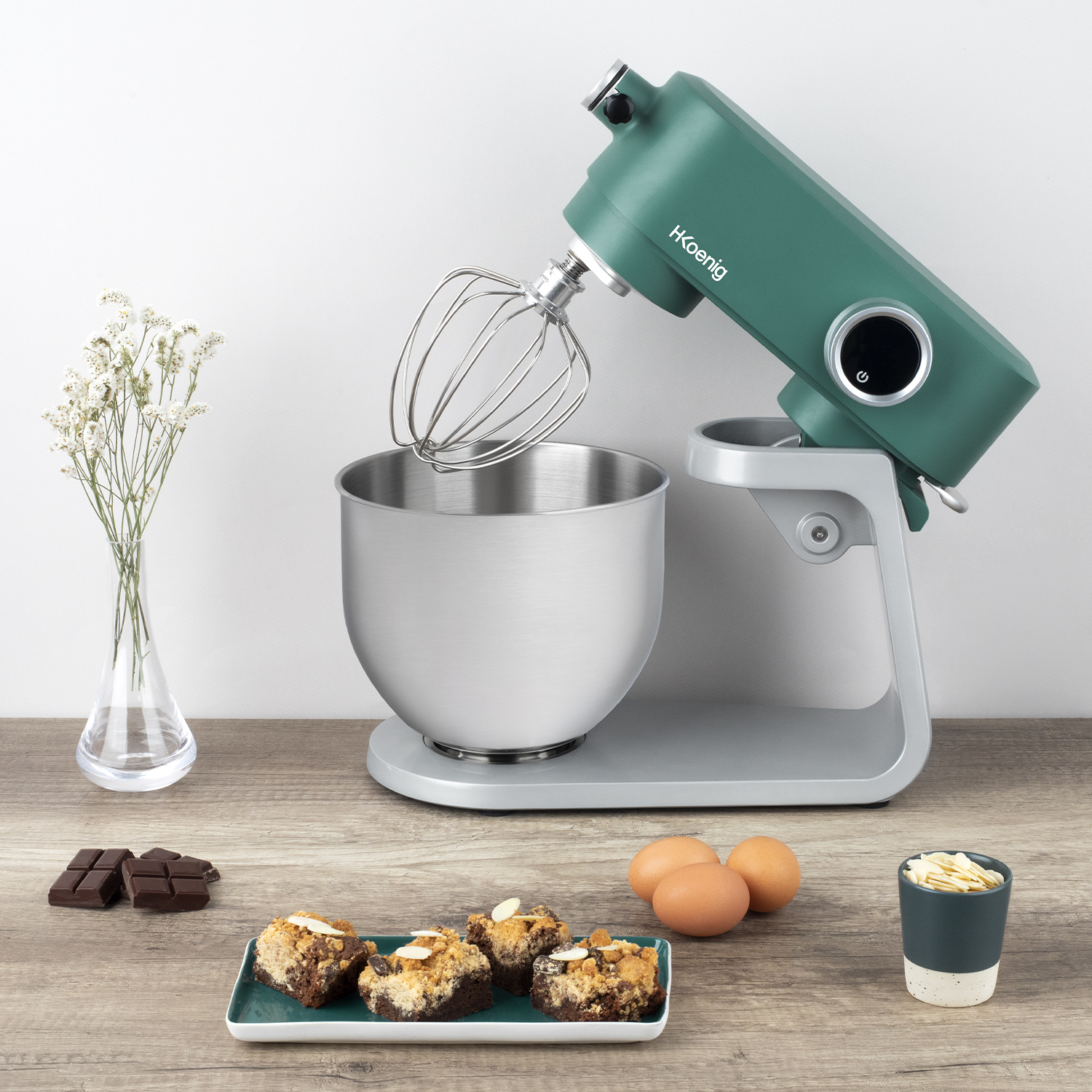 Un'impastatrice verde con una ciotola e accessori, insieme a generi alimentari su una superficie di legno.