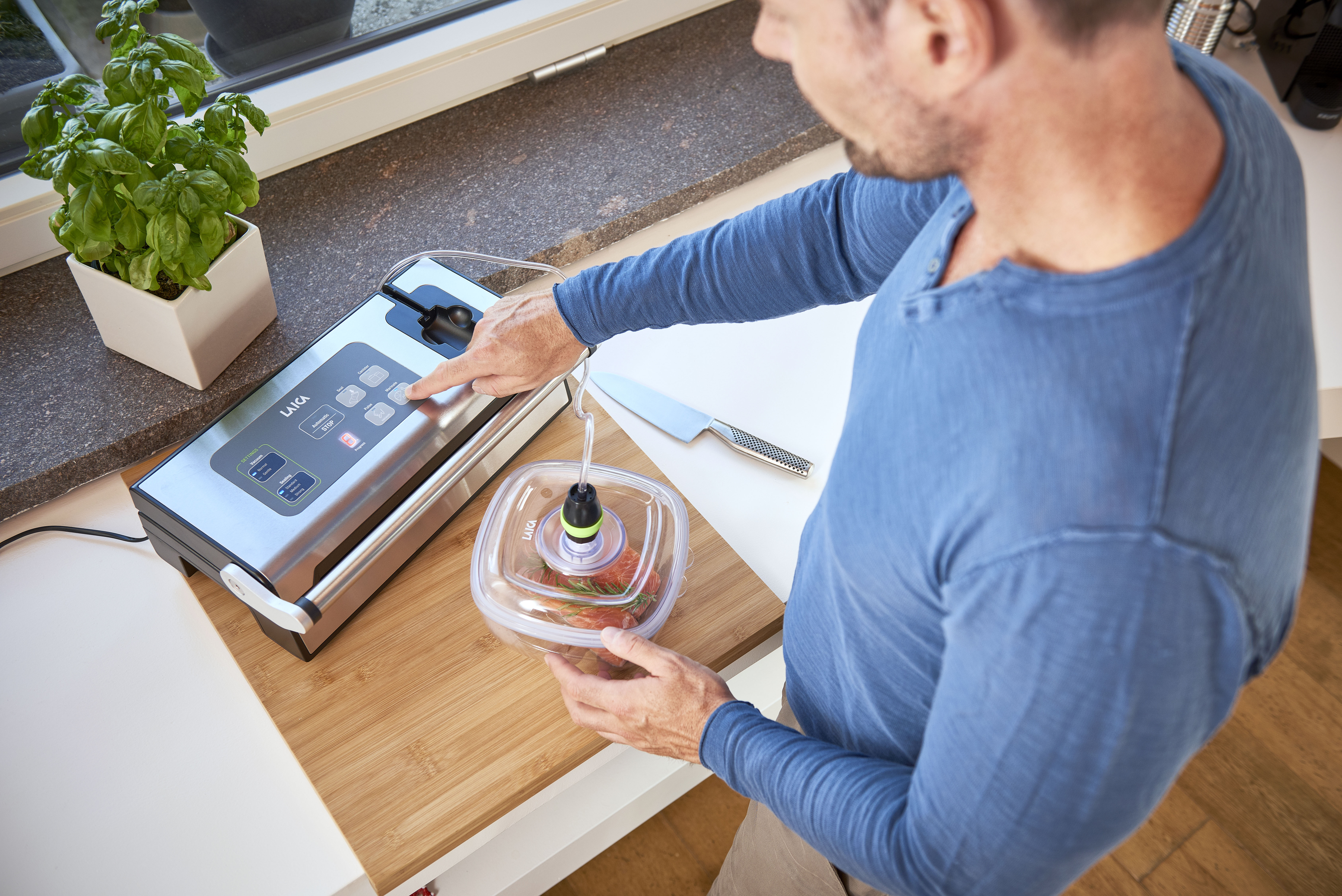 Mężczyzna używa zgrzewarki próżniowej. Jest na drewnianej desce z nożem i bazylią w tle.