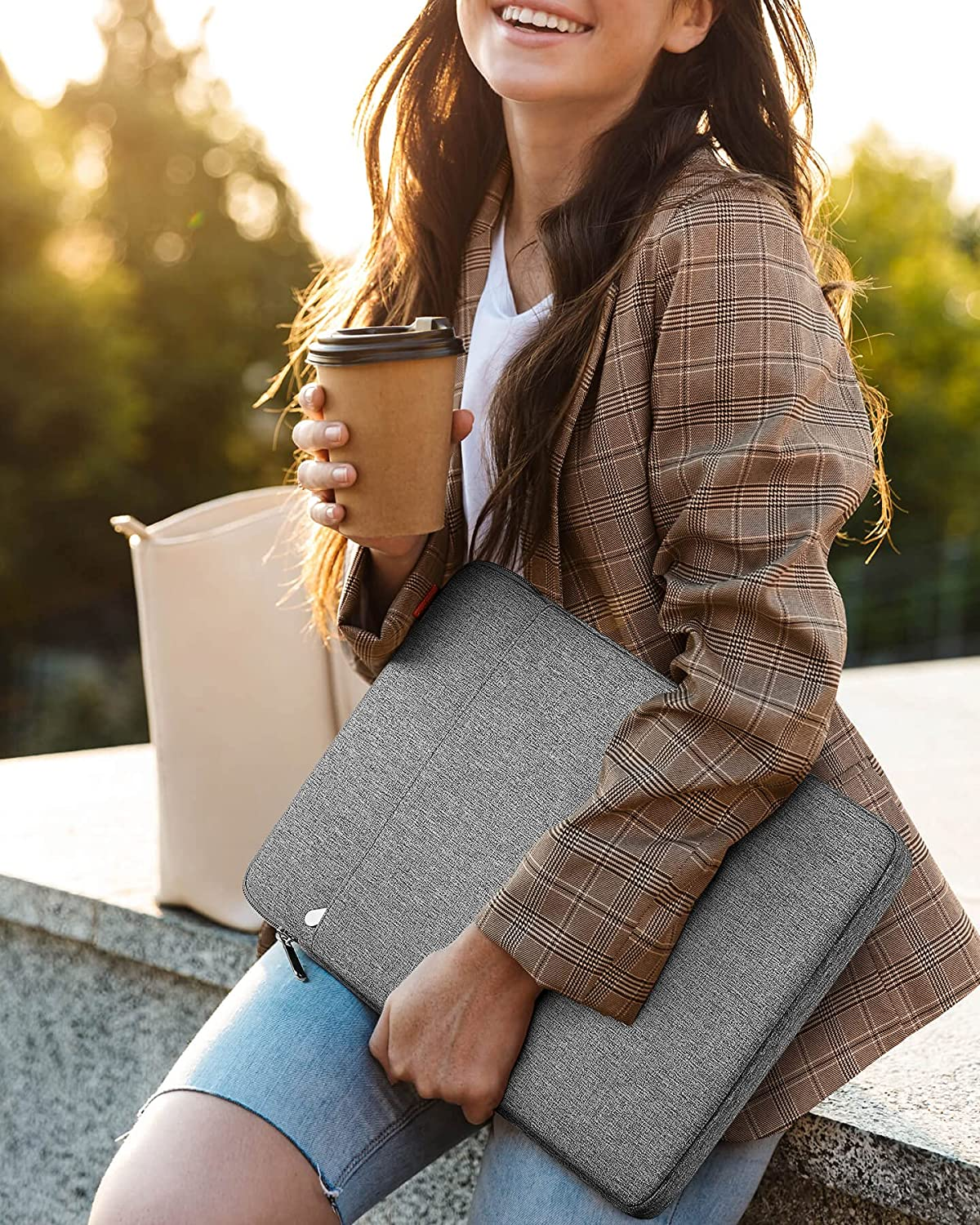 Eine Frau hält eine graue Laptop-Tasche und einen Kaffeebecher im Freien, lächelnd. Sie trägt eine Jacke.
