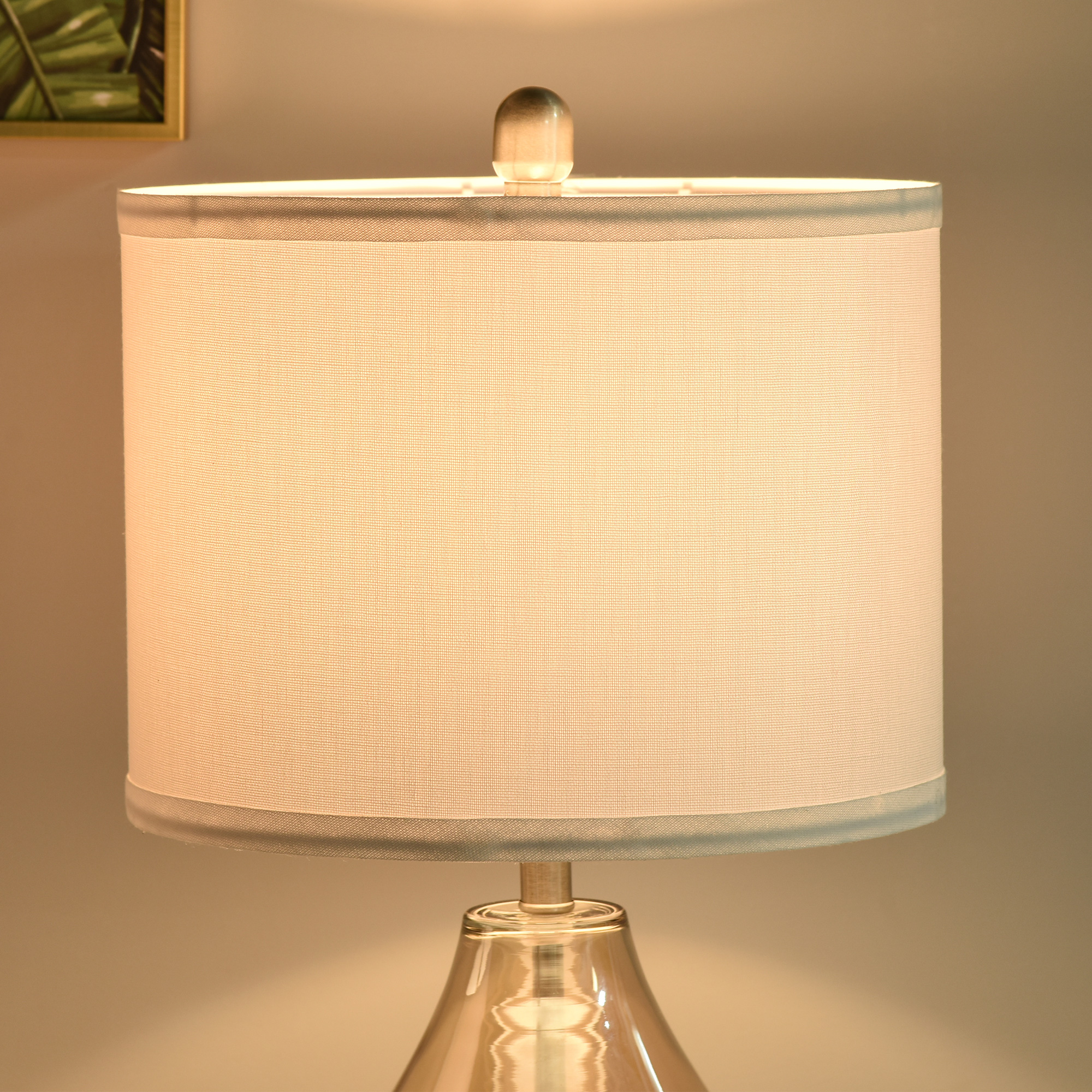 Una lámpara de mesa encendida con pantalla color crema y base de cristal.