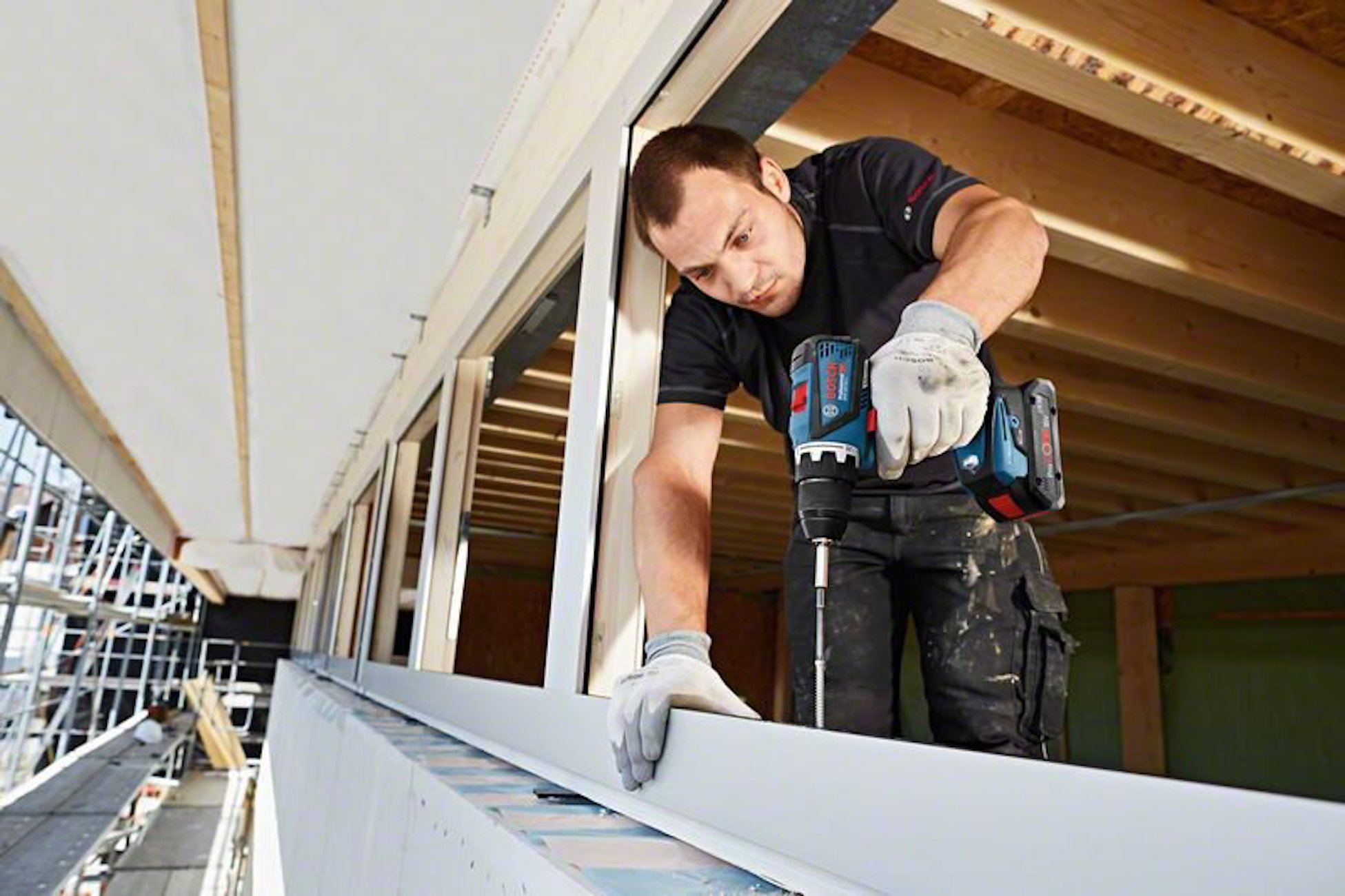 Un uomo usa un trapano elettrico su una struttura in legno. Indossa guanti e occhiali protettivi.