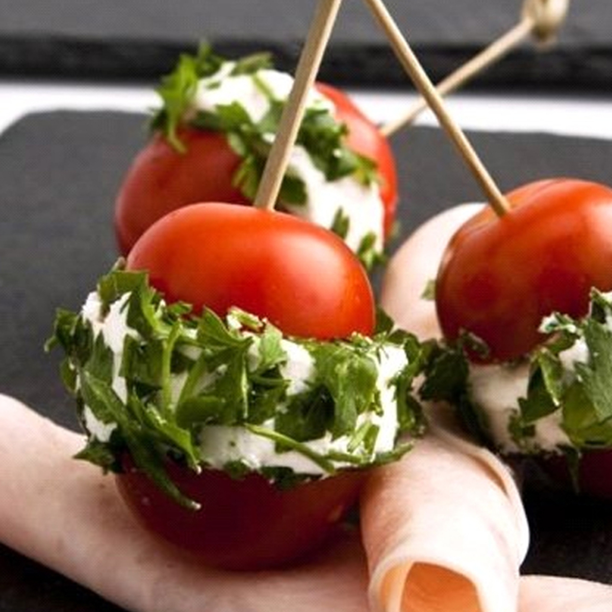 Tomates cubiertos con queso y perejil en brochetas, servidos con jamón sobre una pizarra negra.