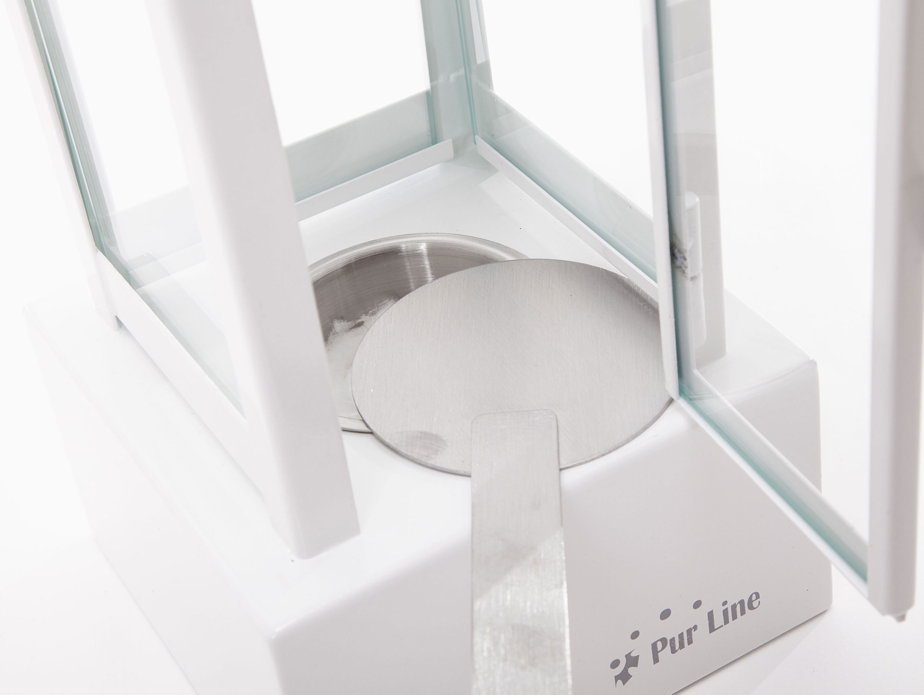 Una balanza de laboratorio blanca. Muestra una placa de metal, una vitrina de vidrio y una espátula de metal.