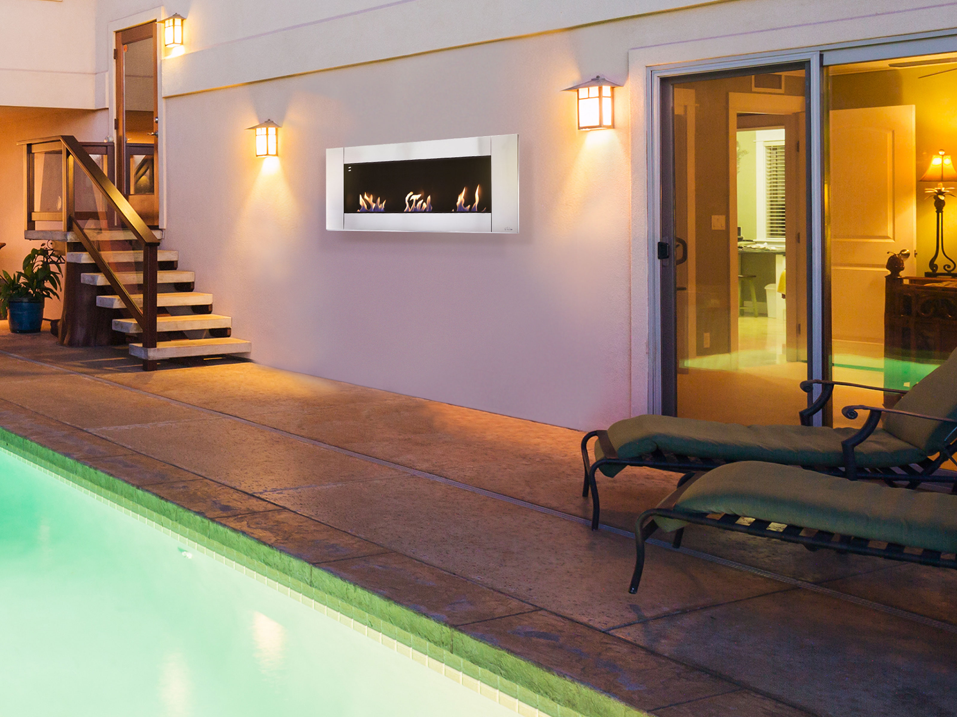 Außenszene bei Nacht. Wandmontierter Kamin mit Flammen. Pool, Liegestühle und Lichter sind sichtbar.