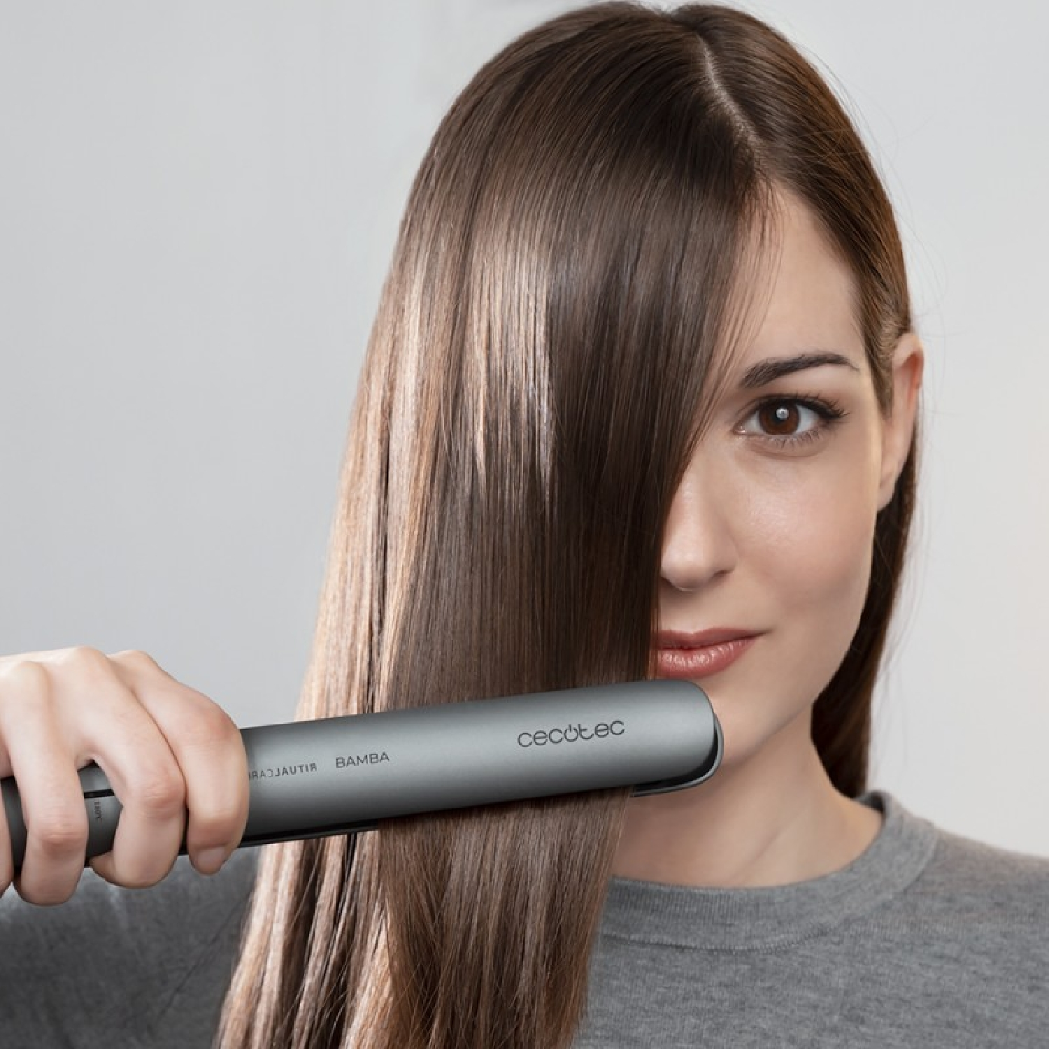Mujer peinándose con plancha. Muestra cabello largo, brillante, castaño y la plancha.