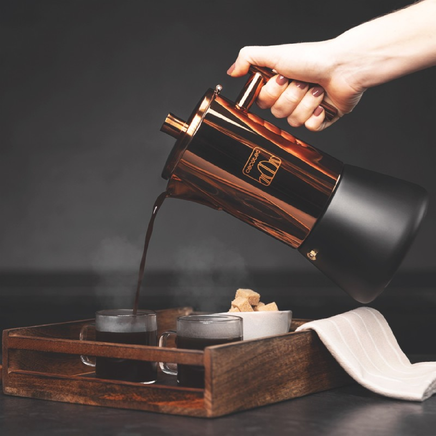 Ręka nalewa kawę z miedzianego garnka do szklanych filiżanek, z kostkami cukru i tacą.