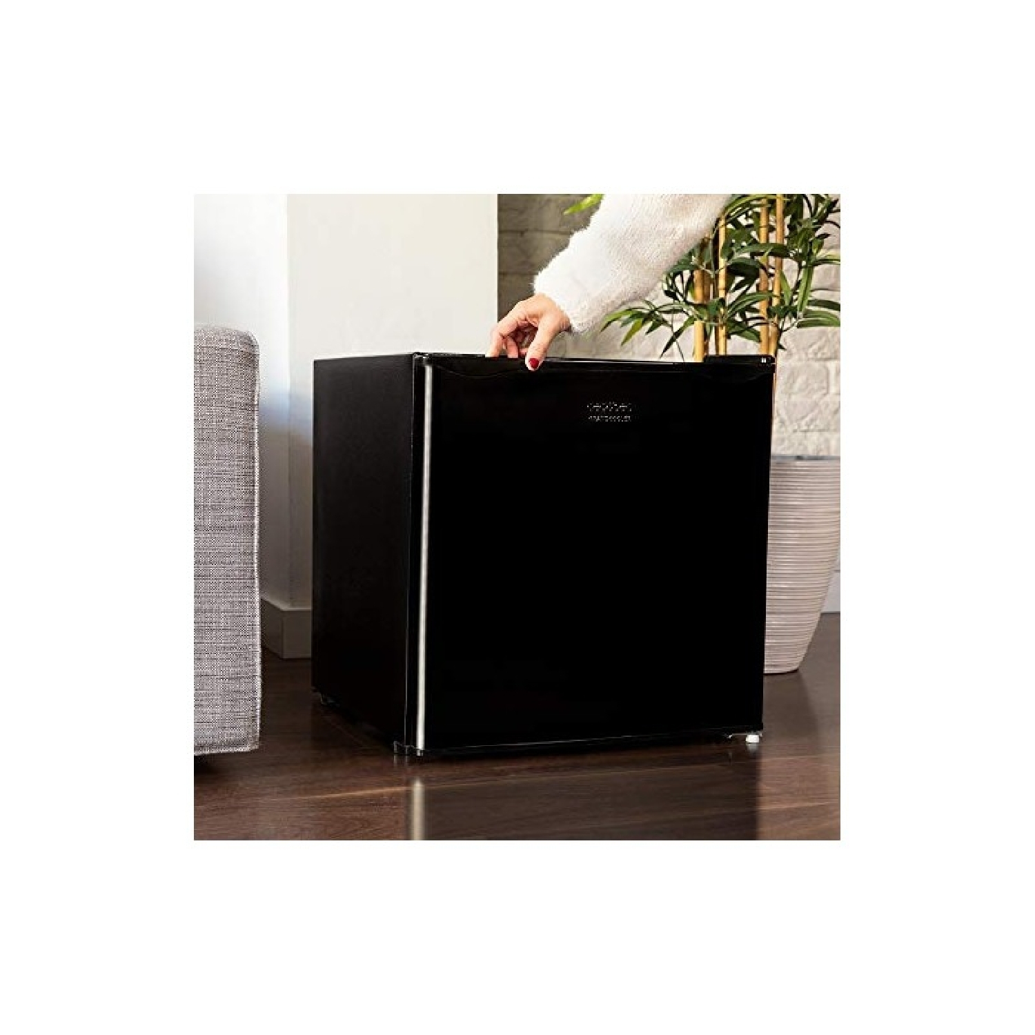 Mini frigo nero con mano sopra, posto su pavimento in legno, vicino a divano grigio e pianta.